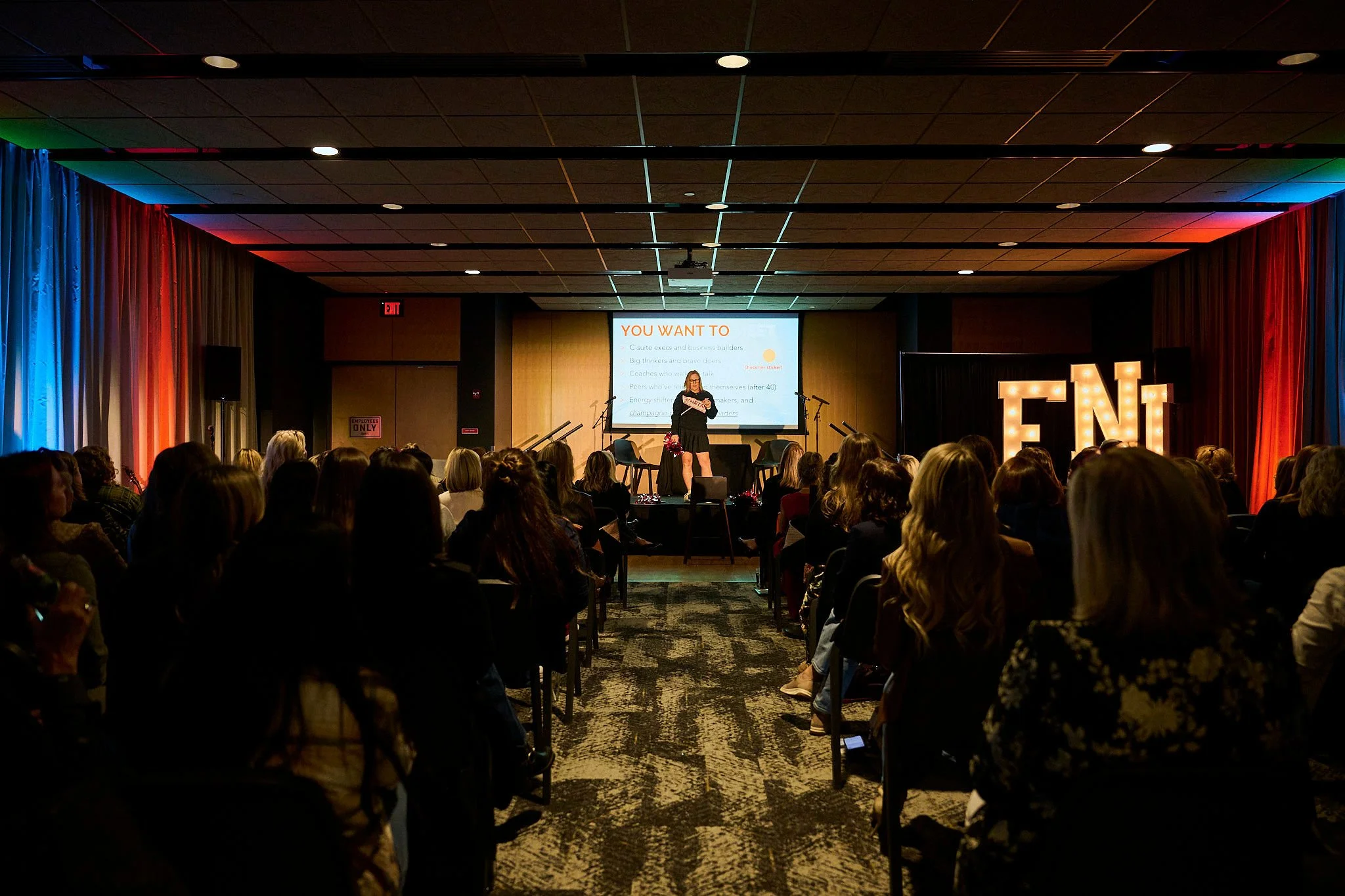 A woman giving a presentation on stage at a conference, with a slide projected behind her and audience members seated facing the stage. Large illuminated letters spelling 'FNL' are on the side of the stage.