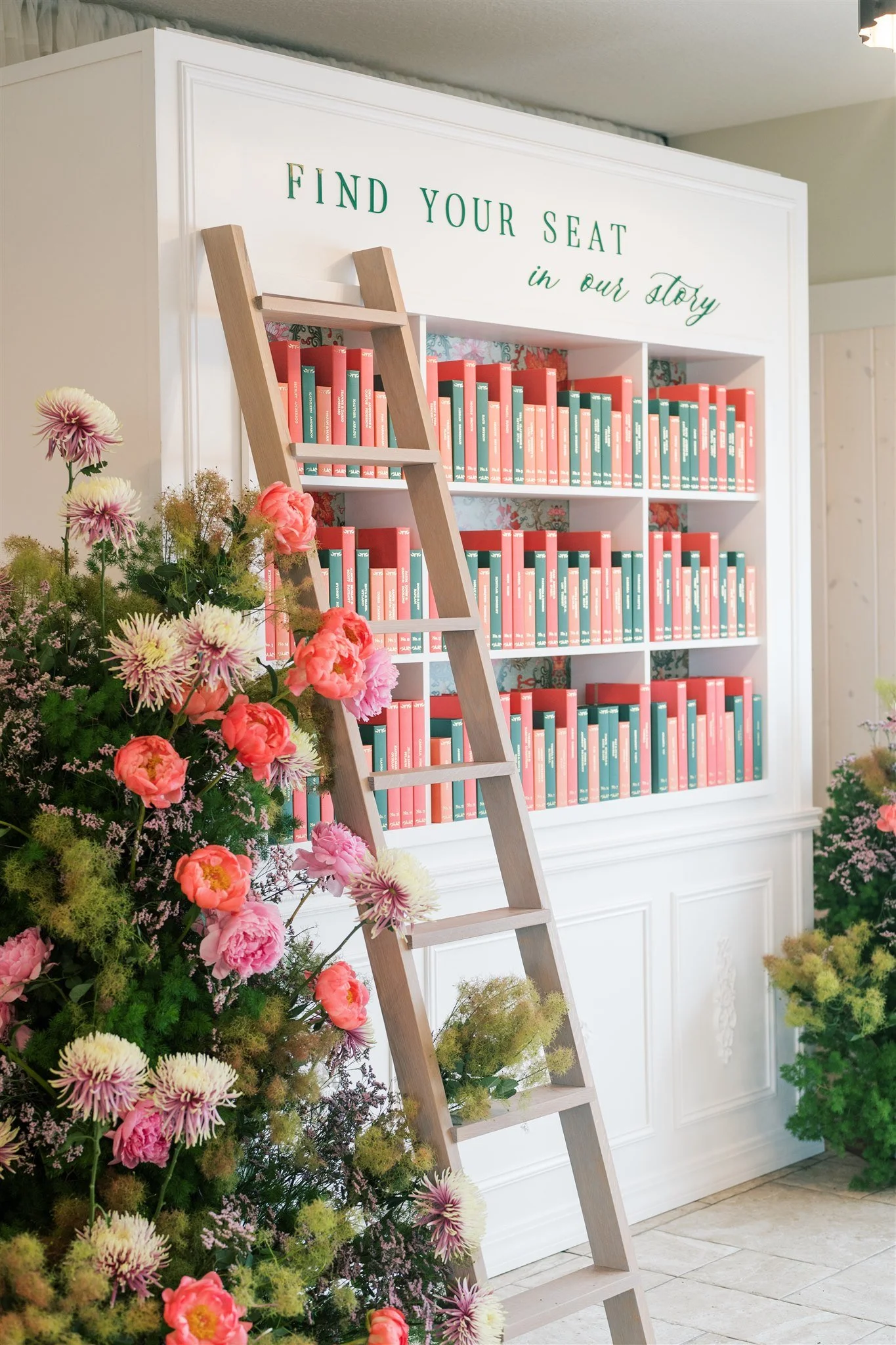 A white bookshelf with the words 'Find Your Seat in our story' on top, filled with red and green books, with a wooden ladder leaning against it. There are colorful flowers, including pink and yellow blossoms, in the foreground and to the side.