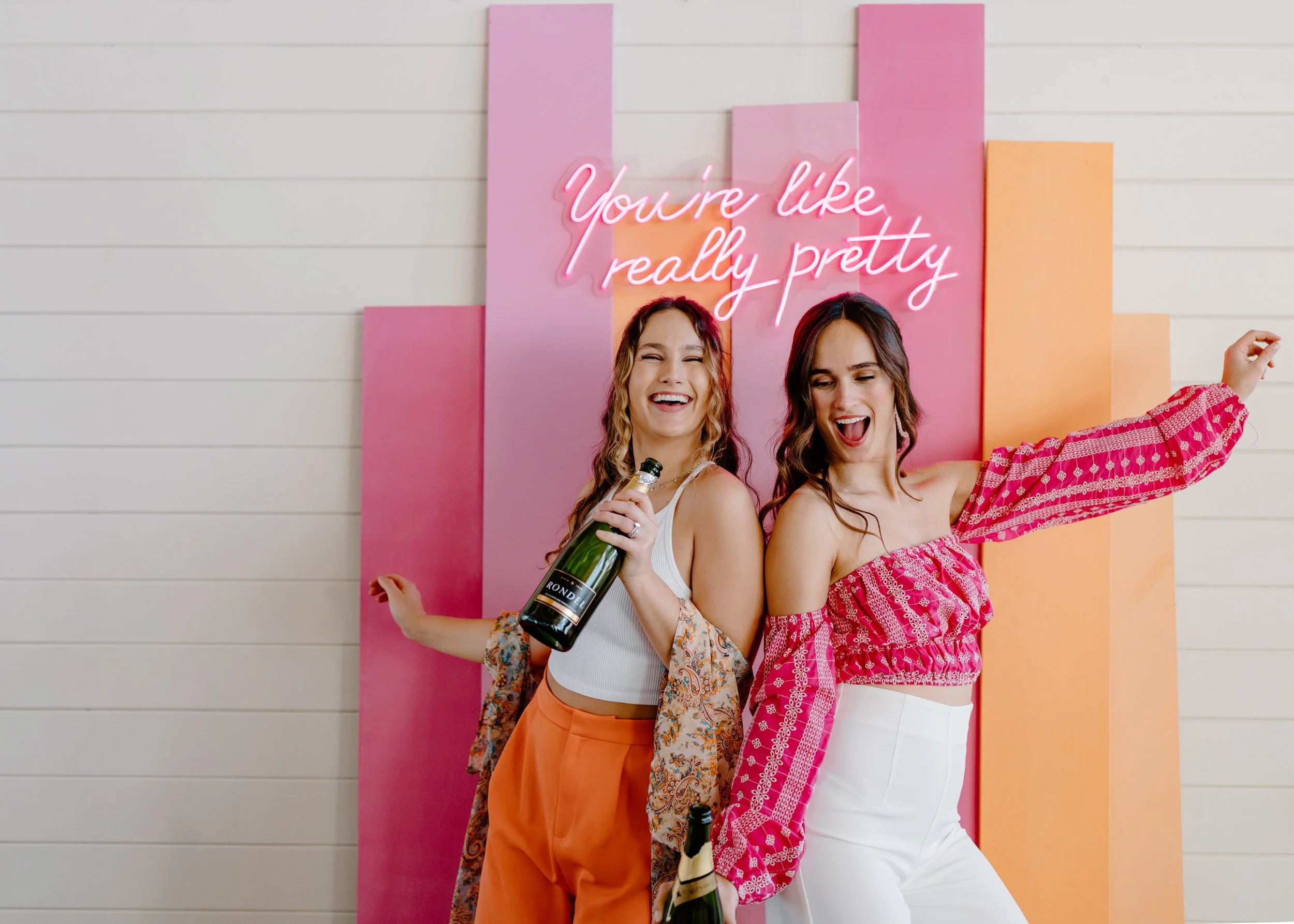 Two young women celebrating with champagne bottles, dancing and smiling in front of a colorful backdrop with a neon sign that reads 'You're like really pretty'.
