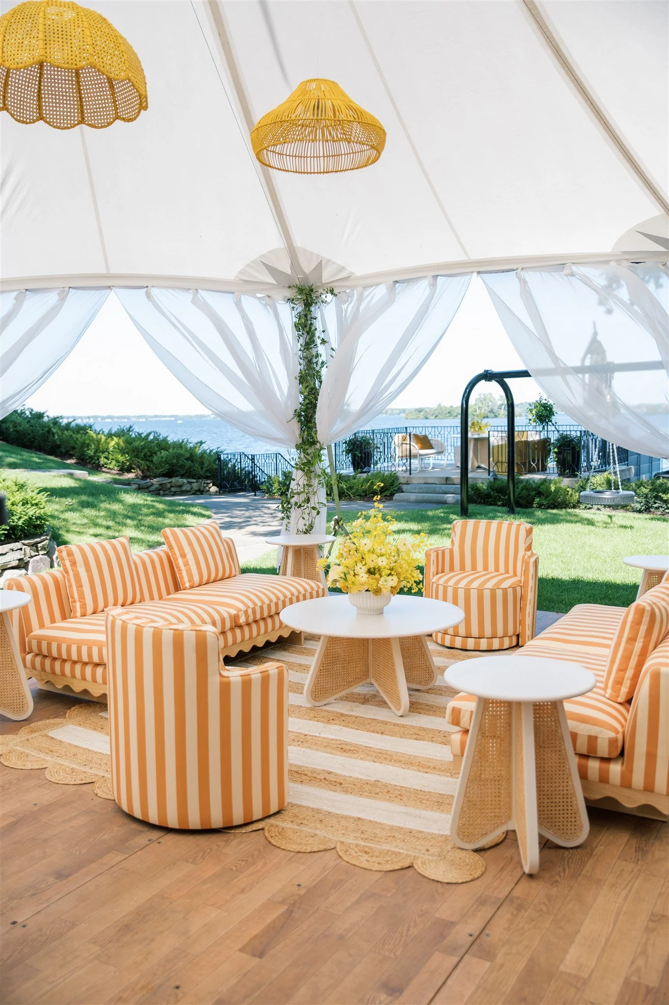 Sunroom outdoor furniture with yellow and white striped cushioned chairs and sofa, white round tables, and a flower arrangement, overlooking a lake with a green lawn and a partially open white canopy.