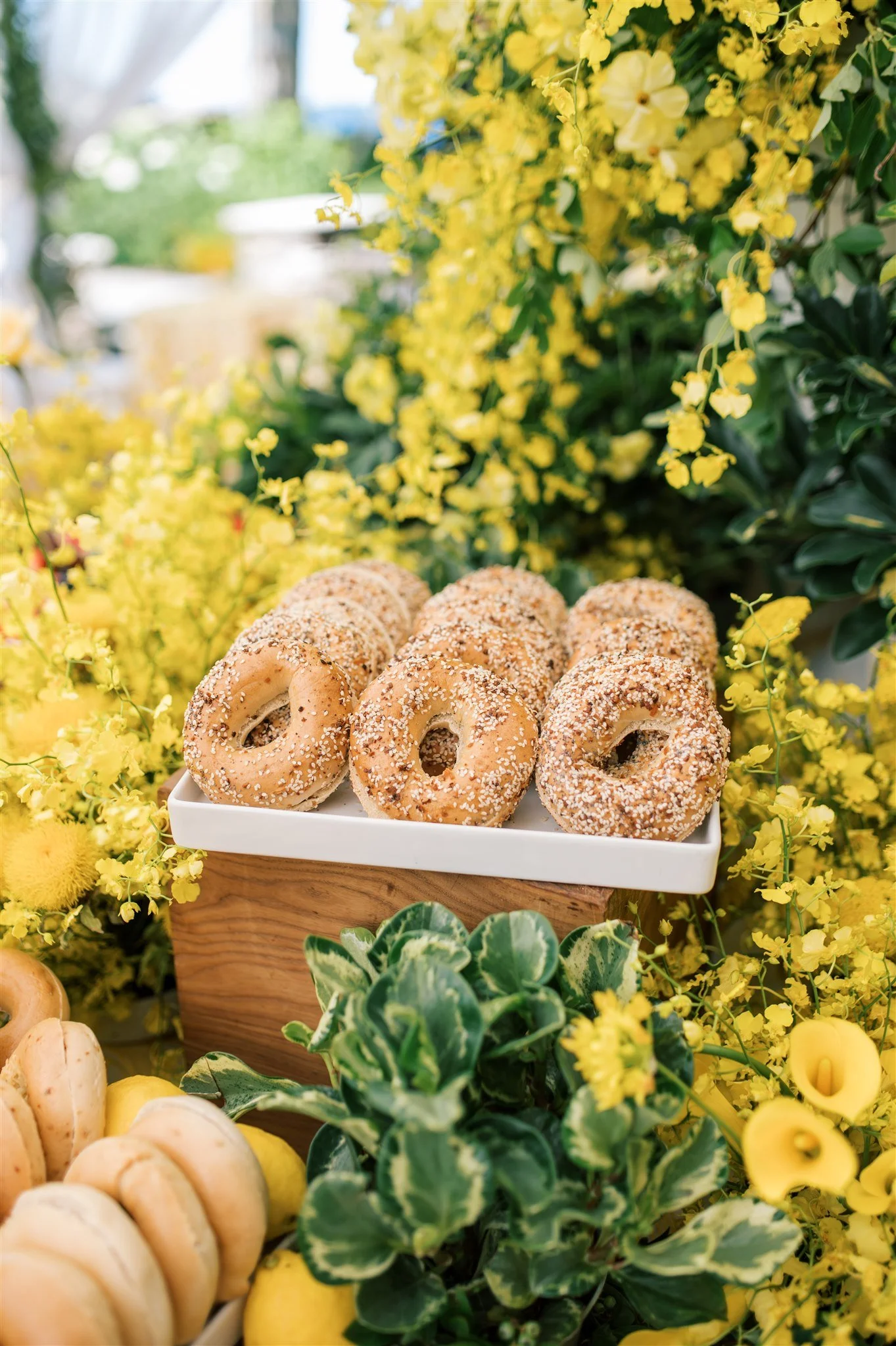 A display of bagels with sesame seeds on a white tray, surrounded by yellow flowers and green foliage.