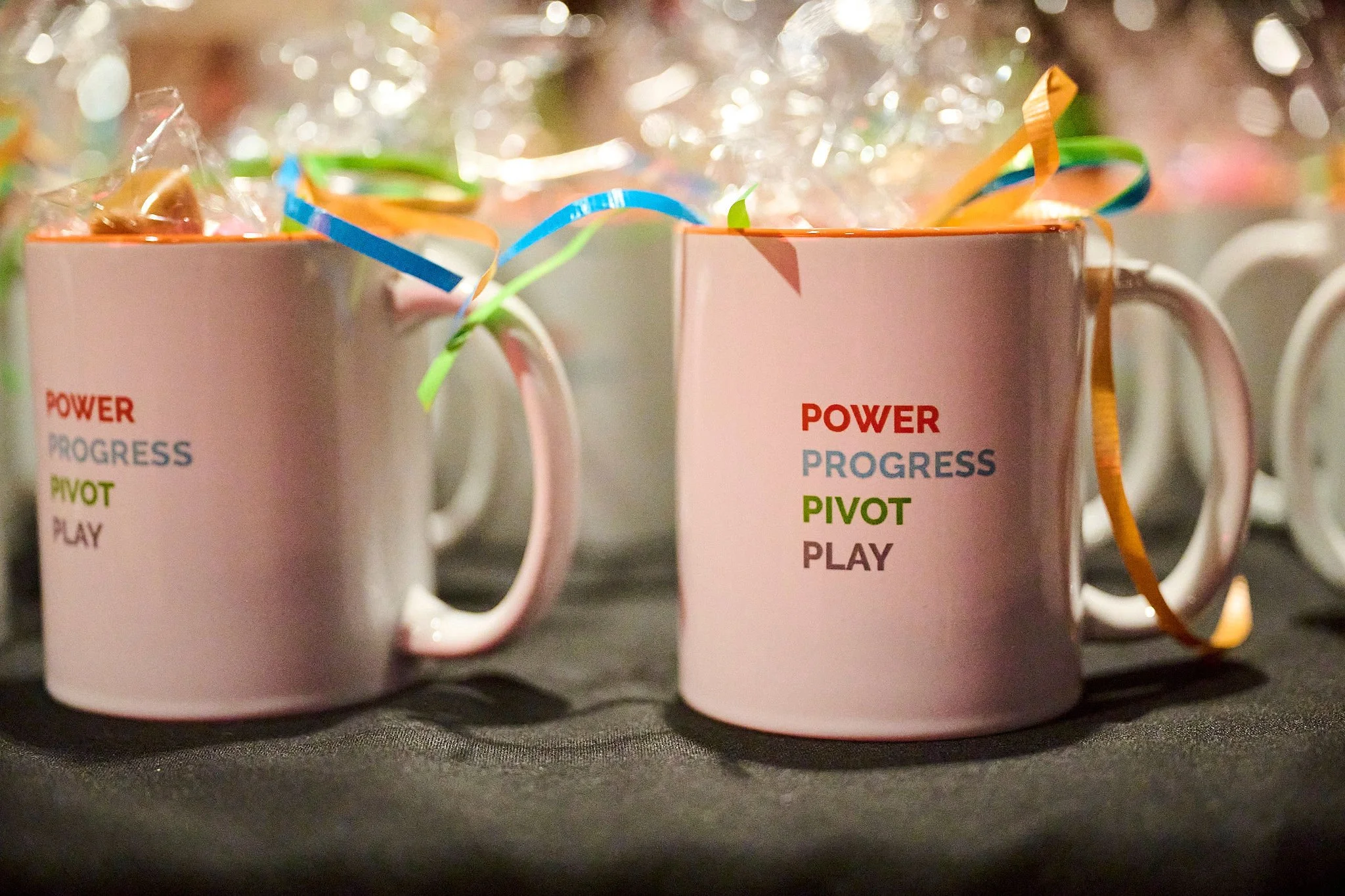 Pink ceramic mugs with colorful listings: 'POWER', 'PROGRESS', 'PIVOT', and 'PLAY' in red, blue, green, and brown text, tied with multicolored curly ribbons, on a dark surface with more mugs in the background.
