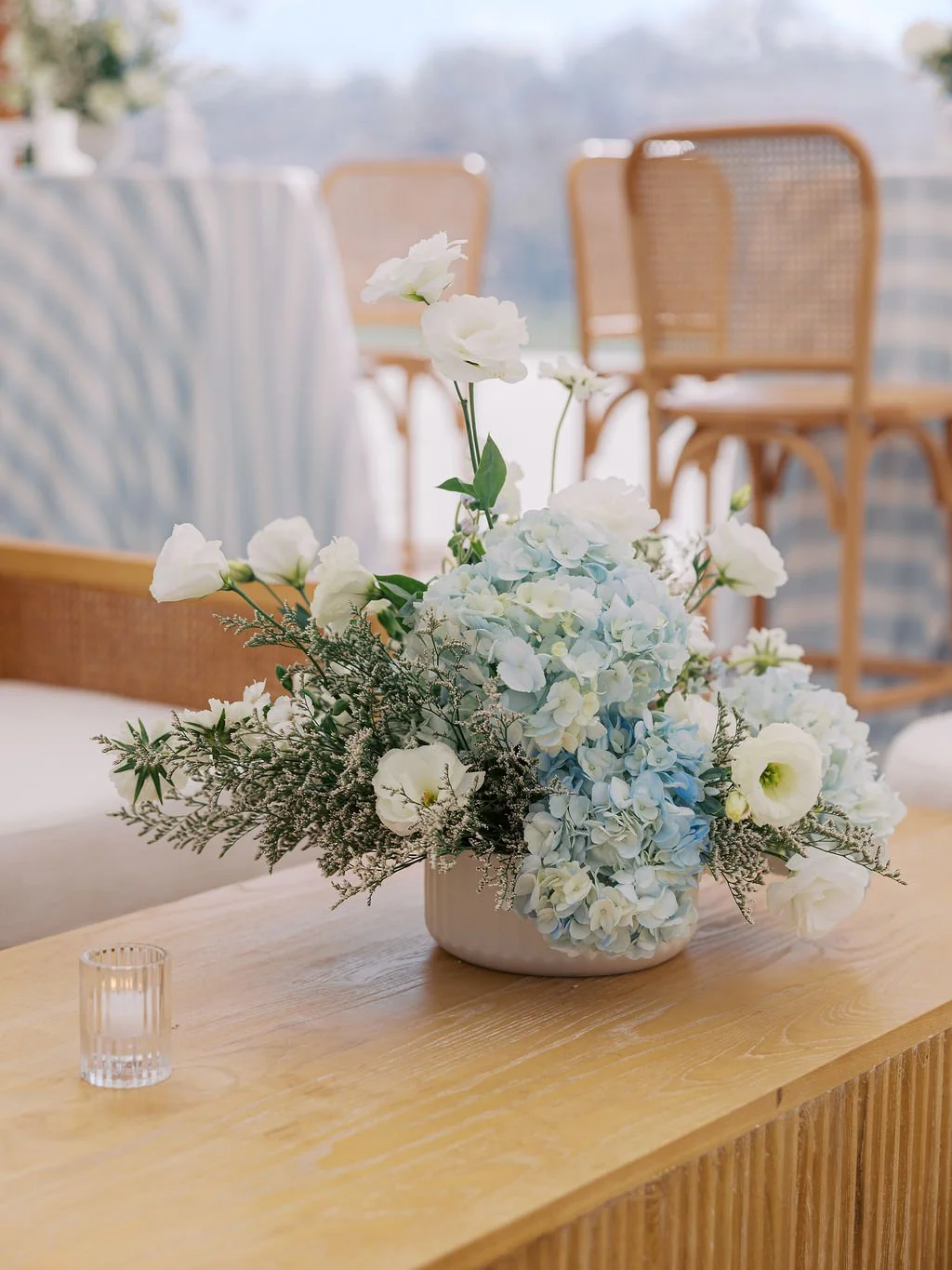 A floral arrangement with white and pale blue hydrangeas, white lisianthus, and greenery in a white vase on a wooden table, with wicker chairs and a patterned tablecloth in the background.