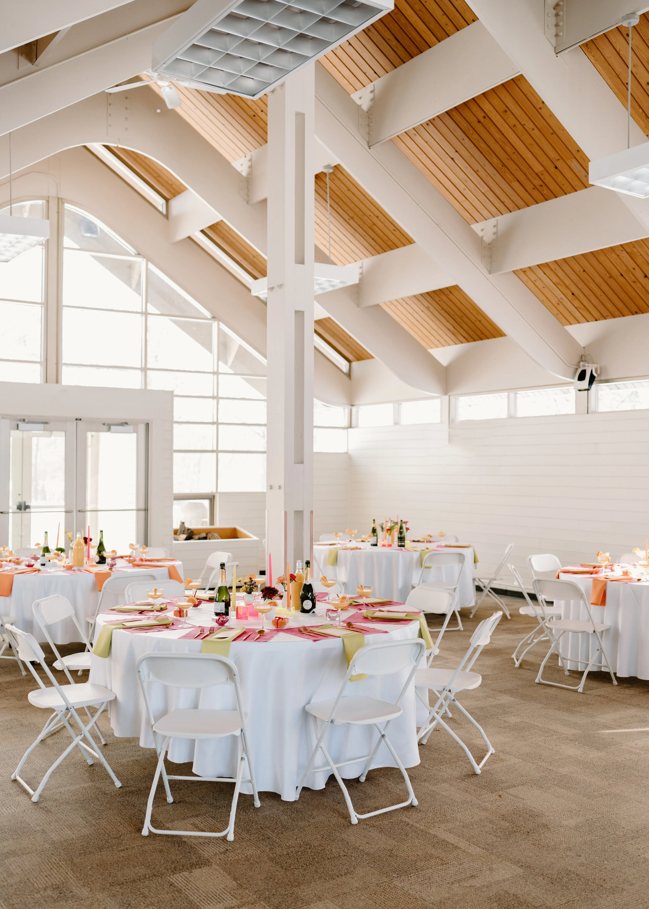 Wedding reception venue with round tables covered in white tablecloths, decorated with pink and green napkins, small floral centerpieces, wine bottles, and lit pink candles. The room has high ceilings with exposed wooden beams and large windows, creating a bright atmosphere.
