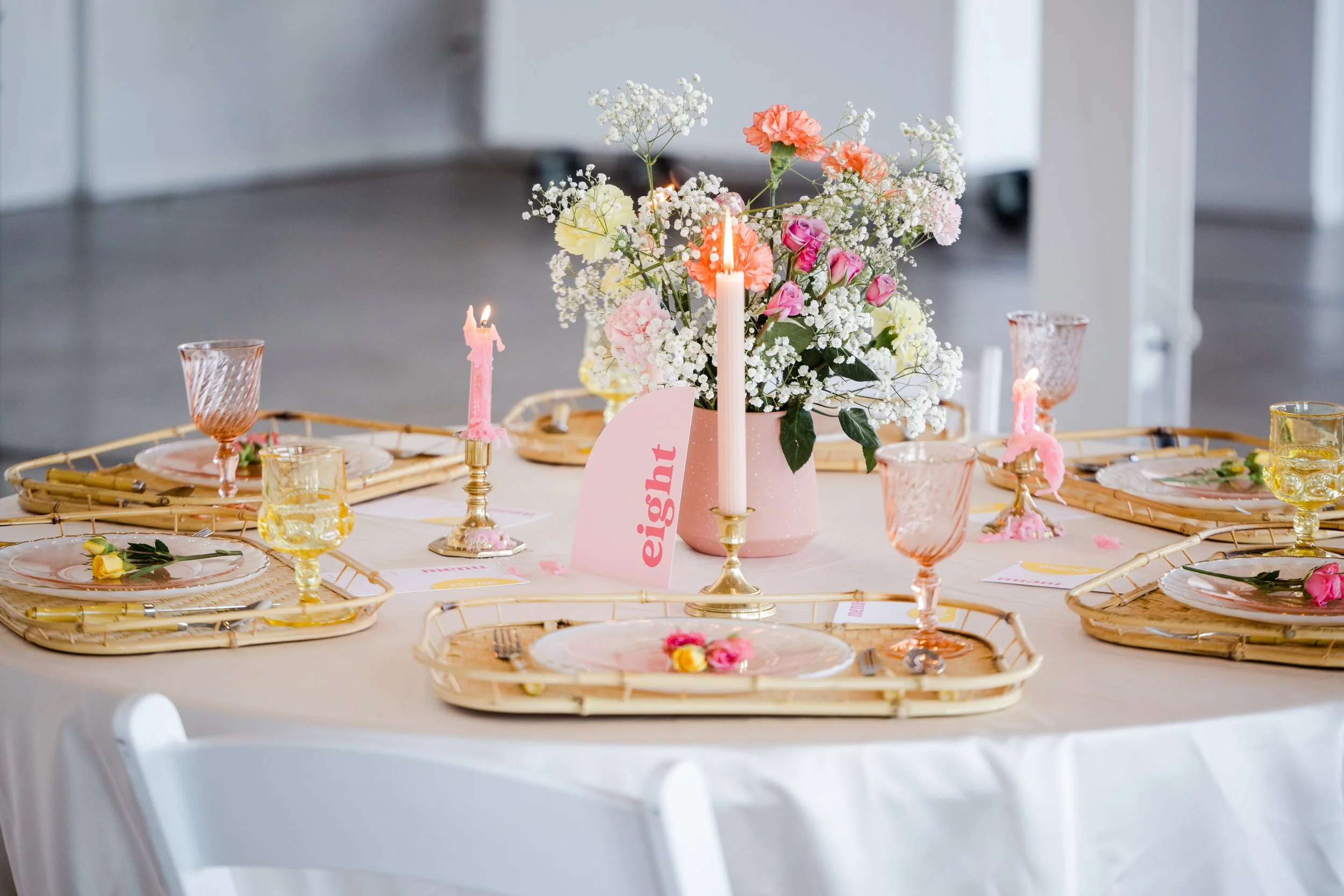 Decorated table with pink and pastel accents, floral centerpiece with pink, white, and yellow flowers, candles, pink glassware, and place settings with pink plates and flowers
