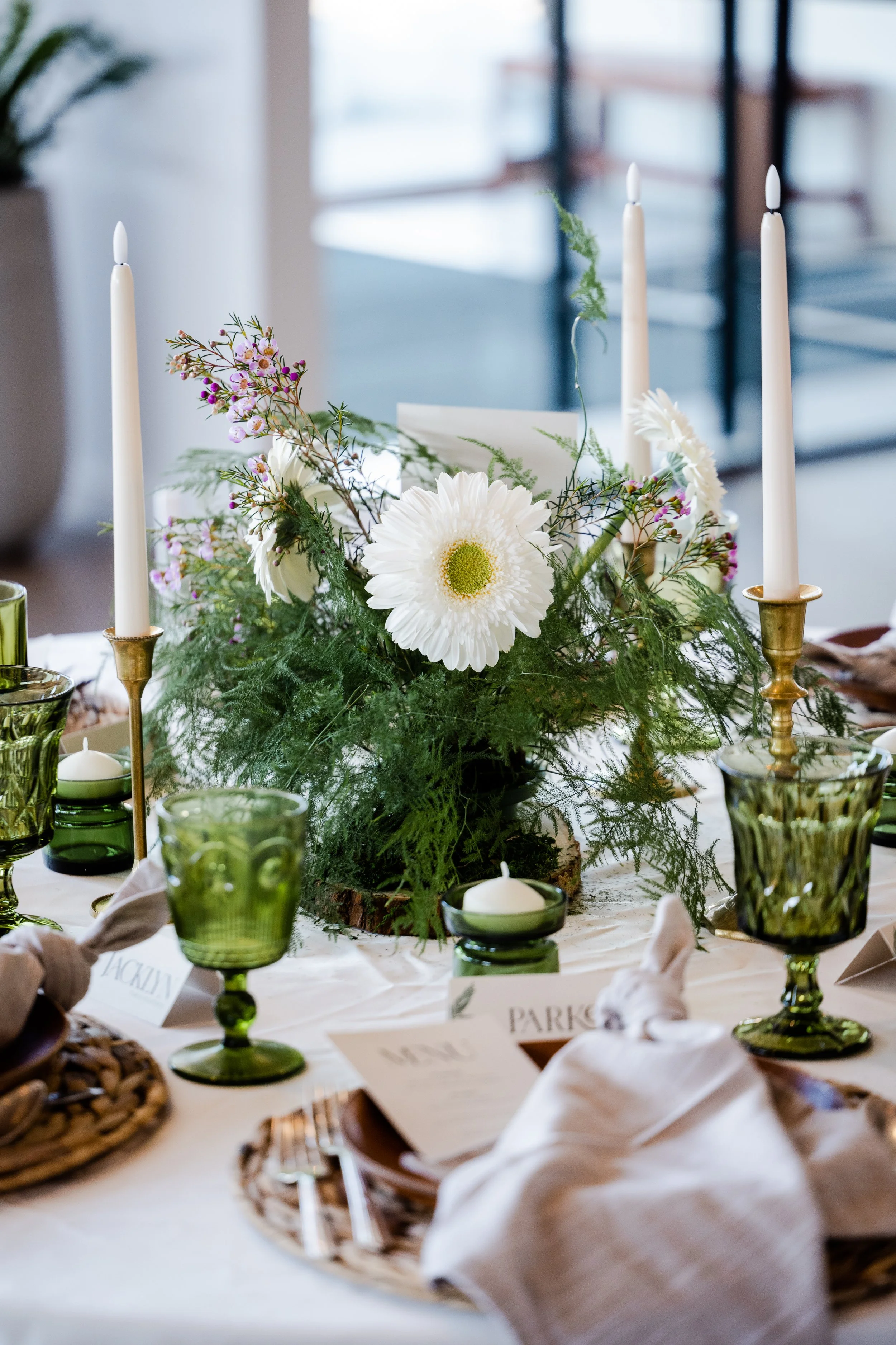 Elegant table centerpiece with white gerbera daisies, pink and purple flowers, green foliage, three white candles, and green glassware, set on a white tablecloth for a formal event.