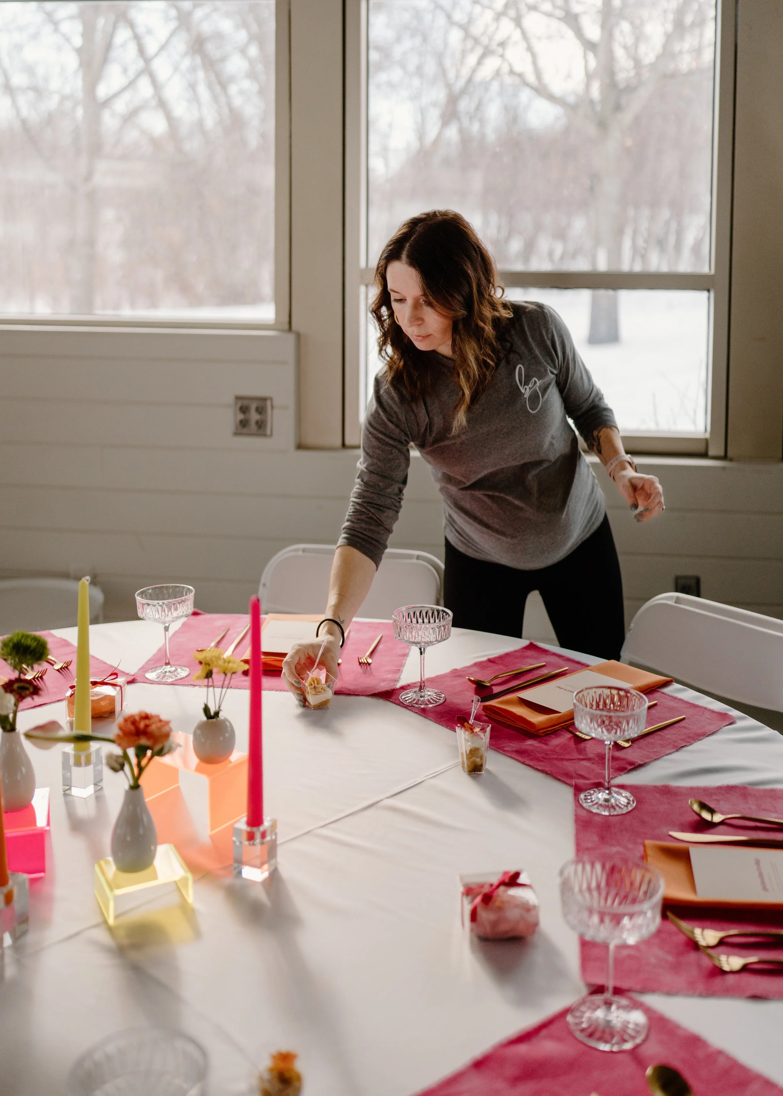 Woman setting a table with pink and orange decorations, candles, and flowers, in a bright room with large windows and snowy landscape outside.