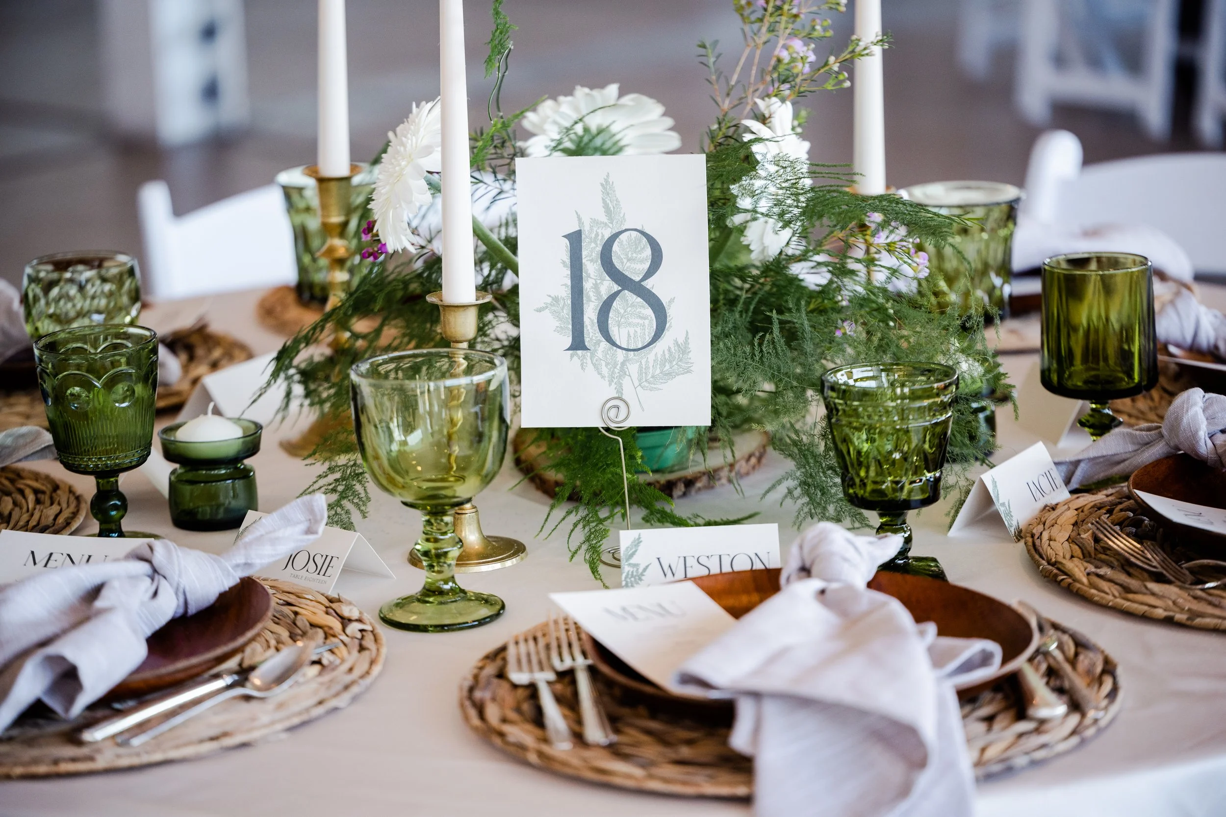 A table set for a formal event with woven placemats, white napkins, green glasses, and a centerpiece of white flowers and greenery. A white card with the number 18 indicates the table number.