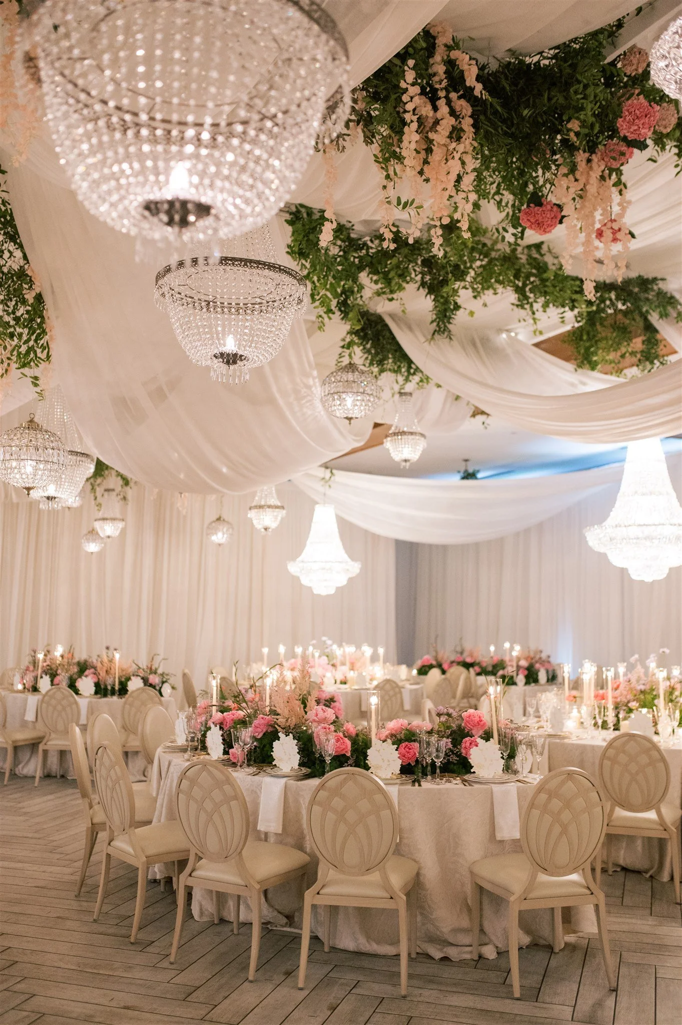 Elegant wedding reception decorated with white drapery, pink and white floral centerpieces, and multiple crystal chandeliers hanging from the ceiling.