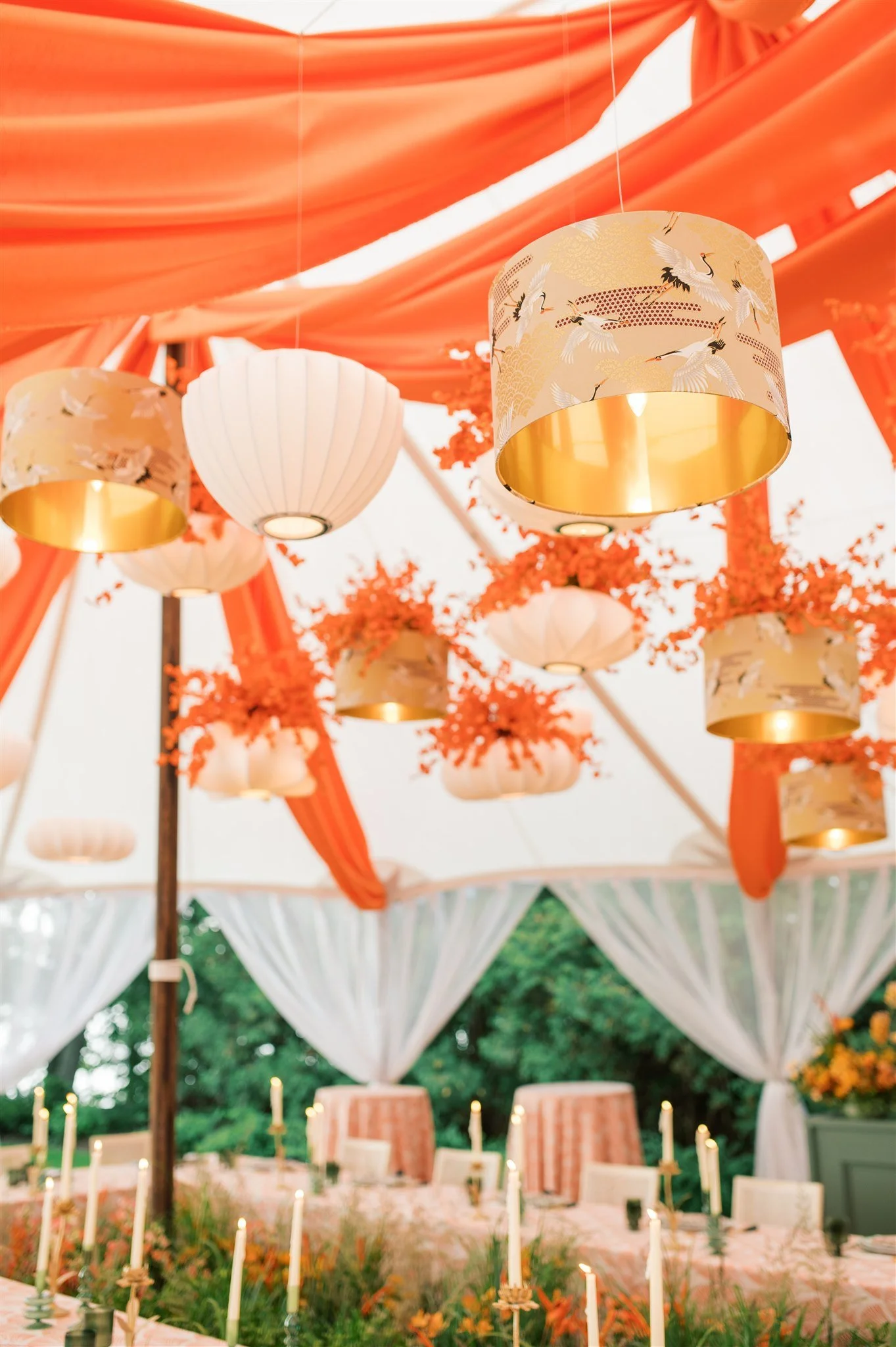 Decorative event tent with orange drapes and hanging lanterns with crane illustrations, white curtains, and a table with candles and floral arrangements inside.