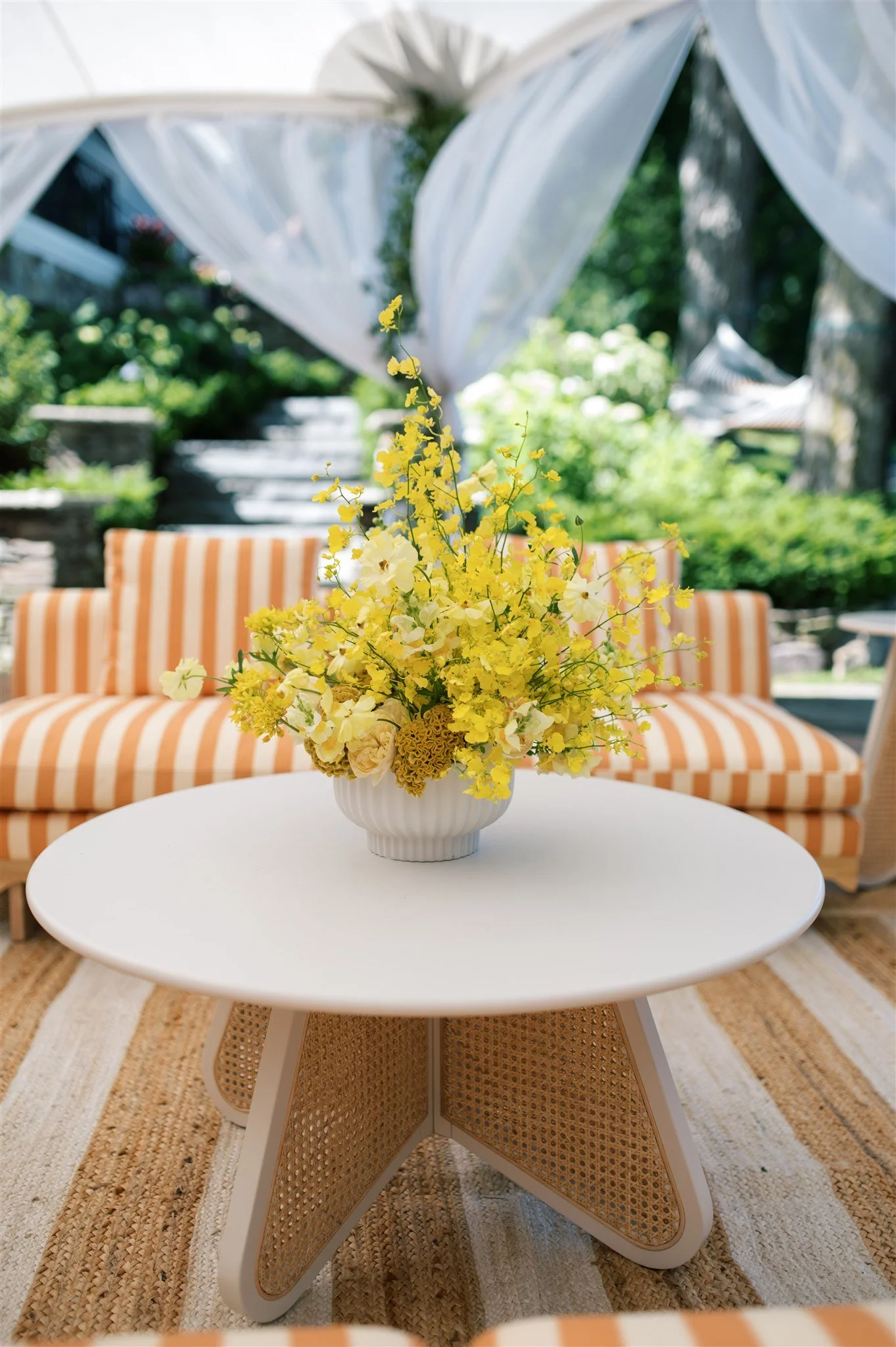 A white round table with a vase of yellow and white flowers, in front of an orange-striped outdoor sofa with cushions, under a sheer white canopy surrounded by greenery.