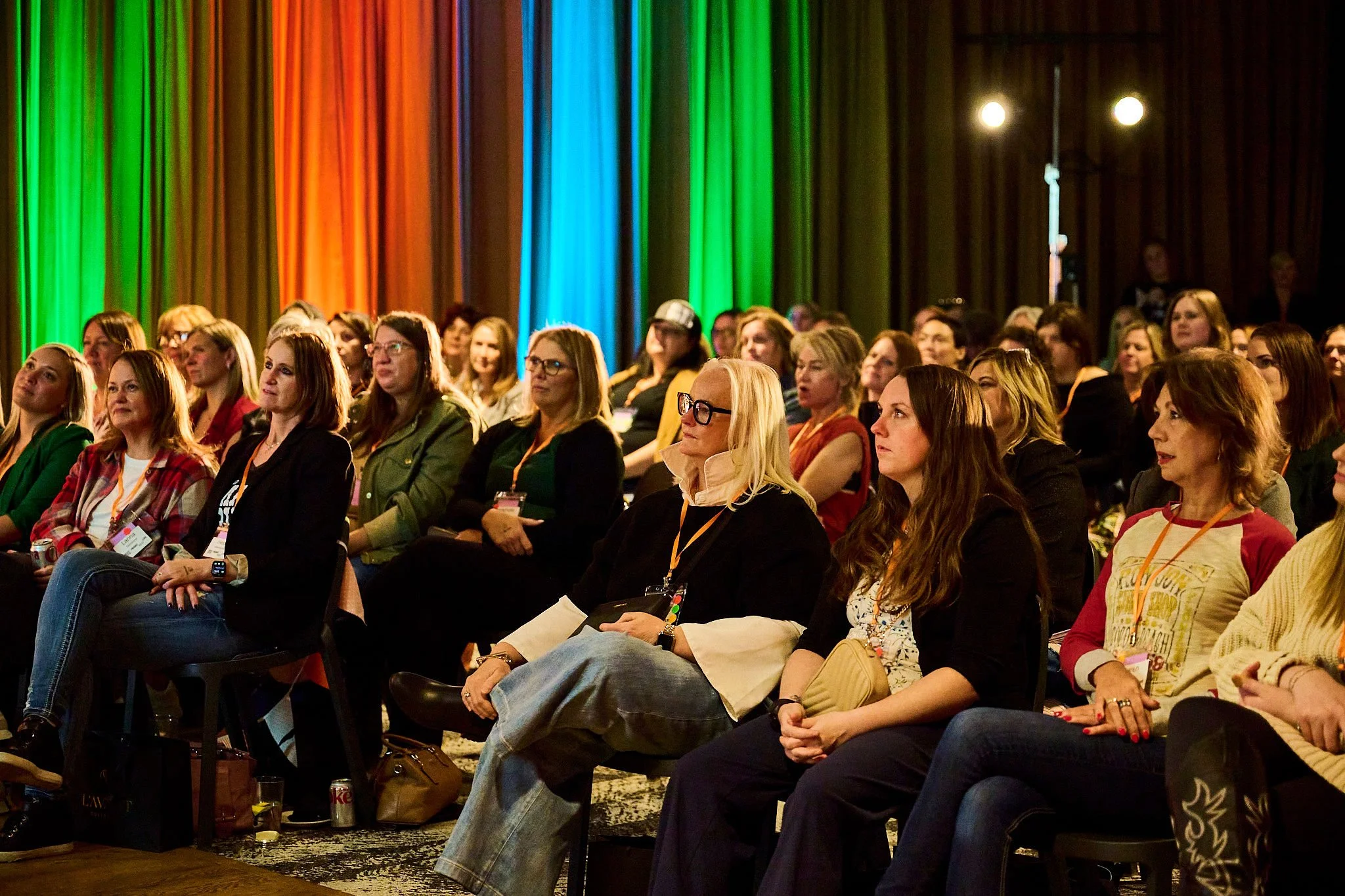 Audience of women watching a presentation or performance in a conference hall with colorful stage curtains in the background.