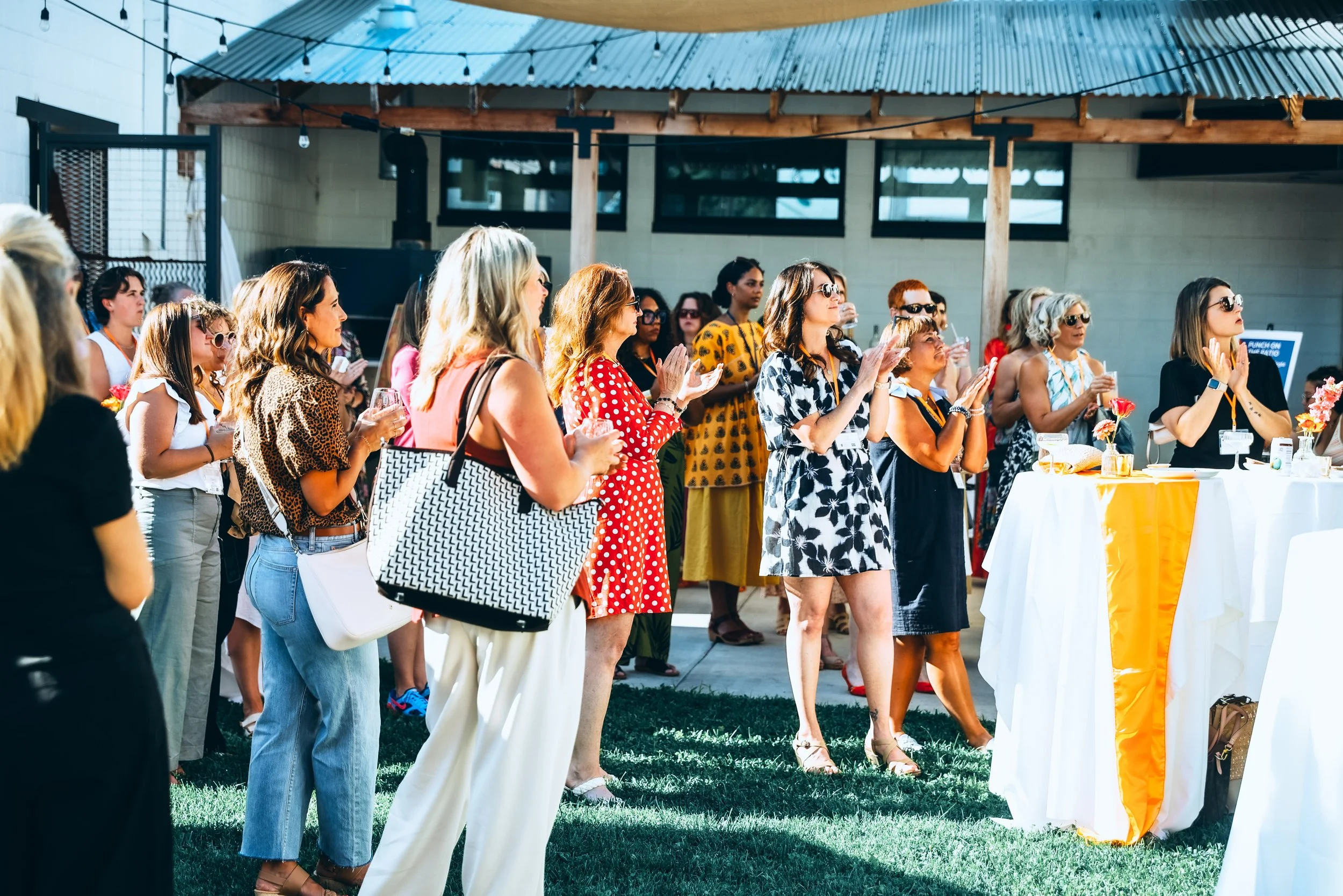 Group of women gathered outdoors, some holding drinks, attending an event on a sunny day with tables decorated in yellow and white.
