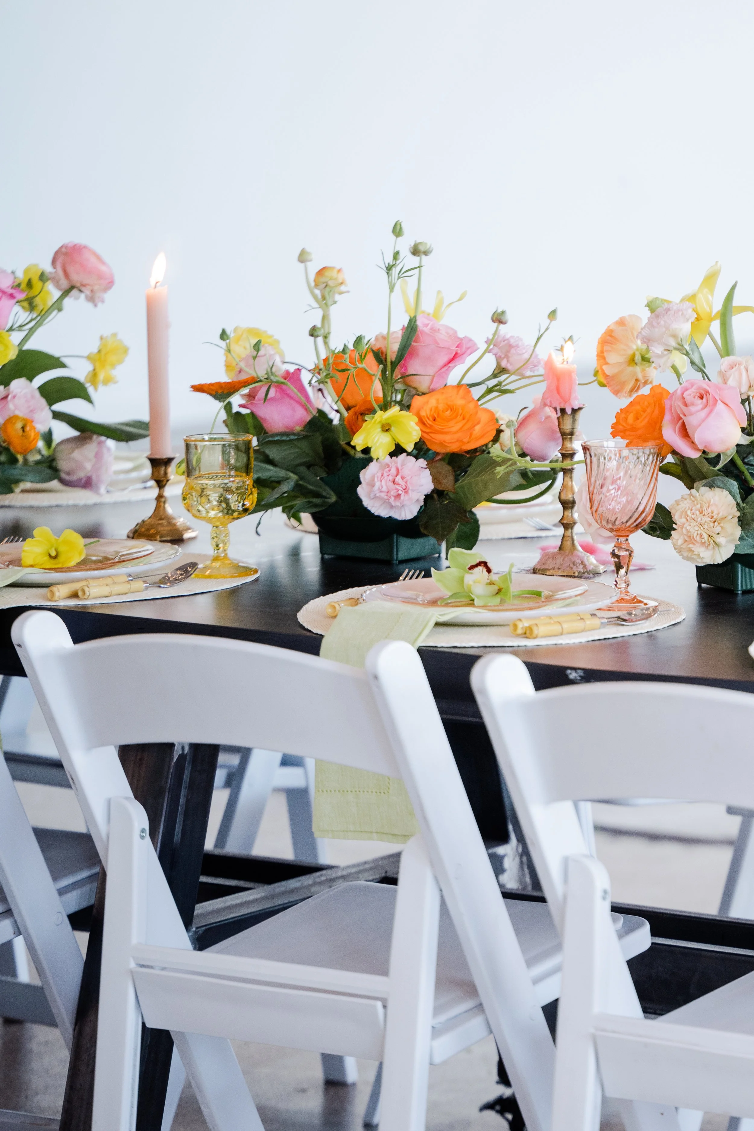 Elegant dining table set with floral centerpiece, pink and orange flowers, pink and yellow candles, and pastel-colored glassware and plates.
