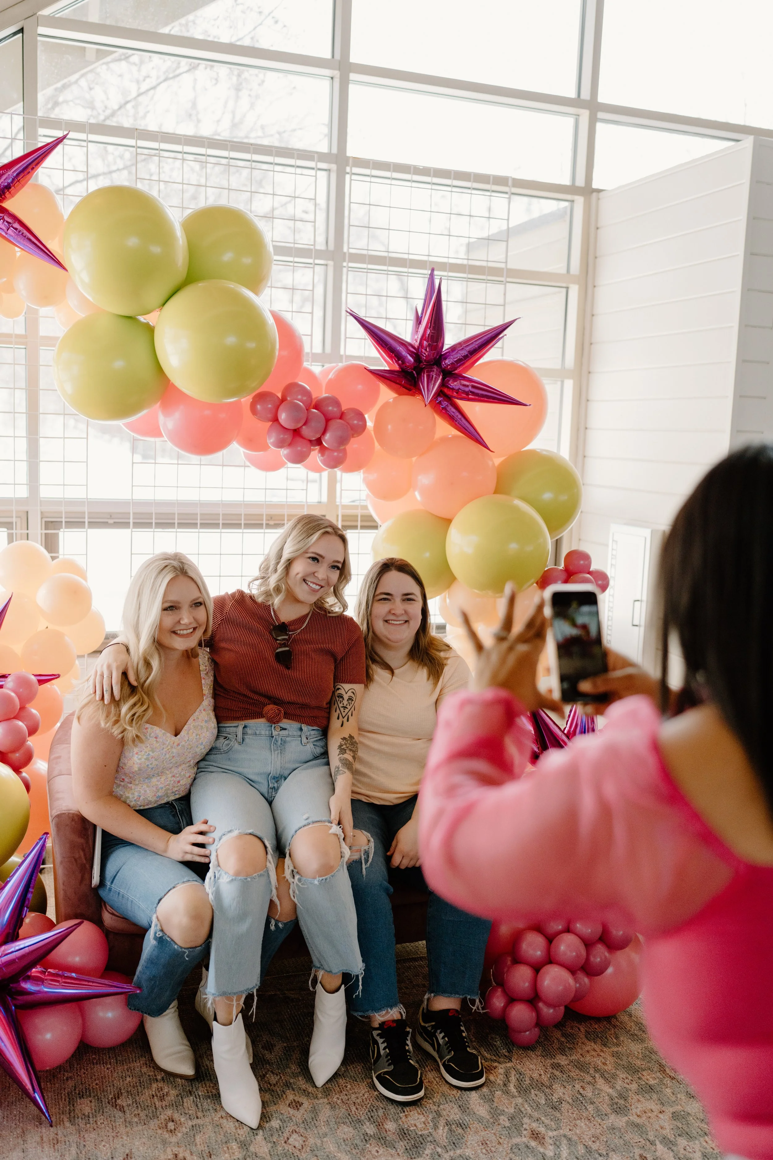 Three women sitting on a sofa, smiling for a photo taken by a woman in pink in front of colorful balloon decorations with stars, pink, and green balloons, in a bright indoor space.