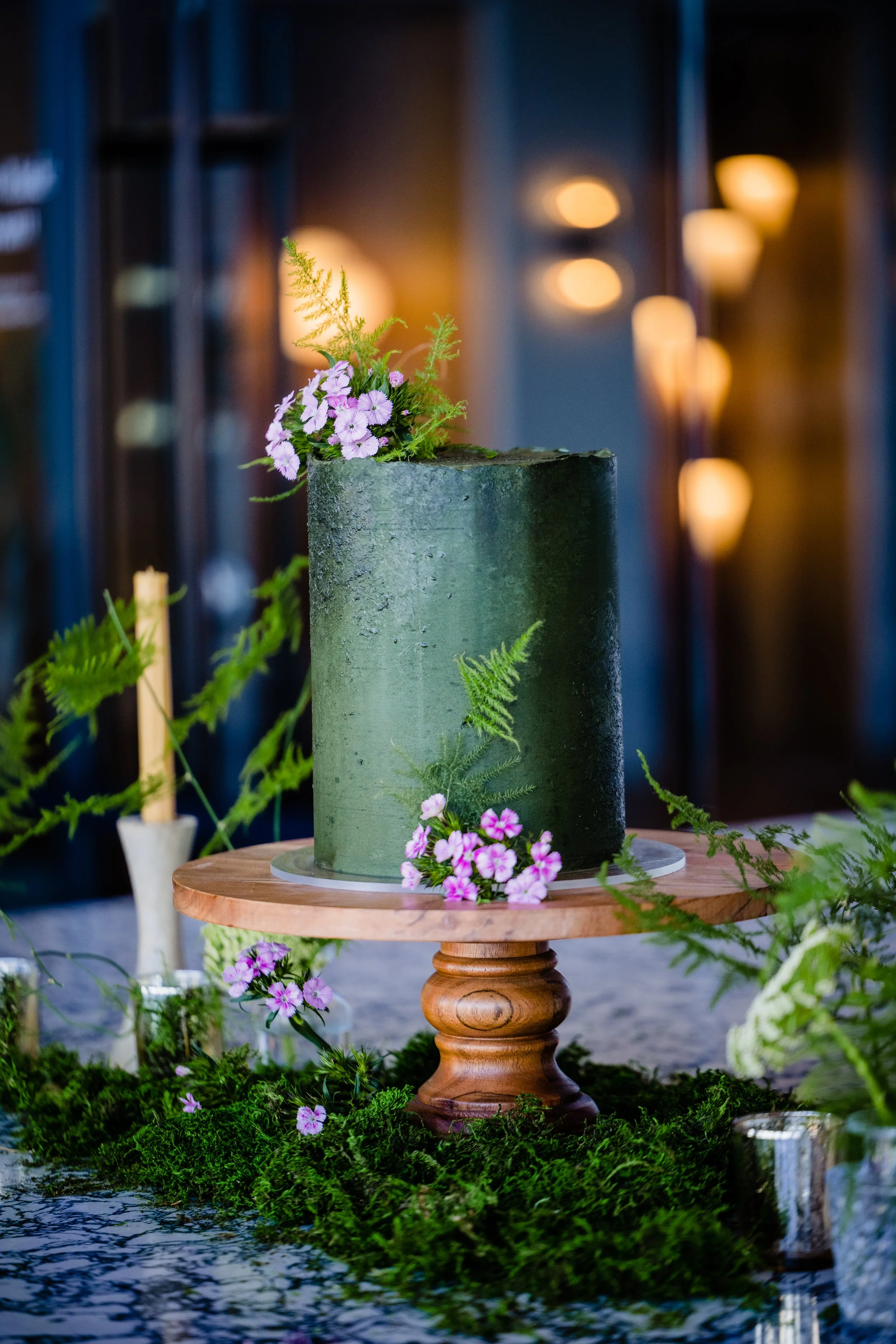 A tall green cake decorated with purple and pink flowers and green ferns, placed on a wooden cake stand amidst green moss and floral arrangements.