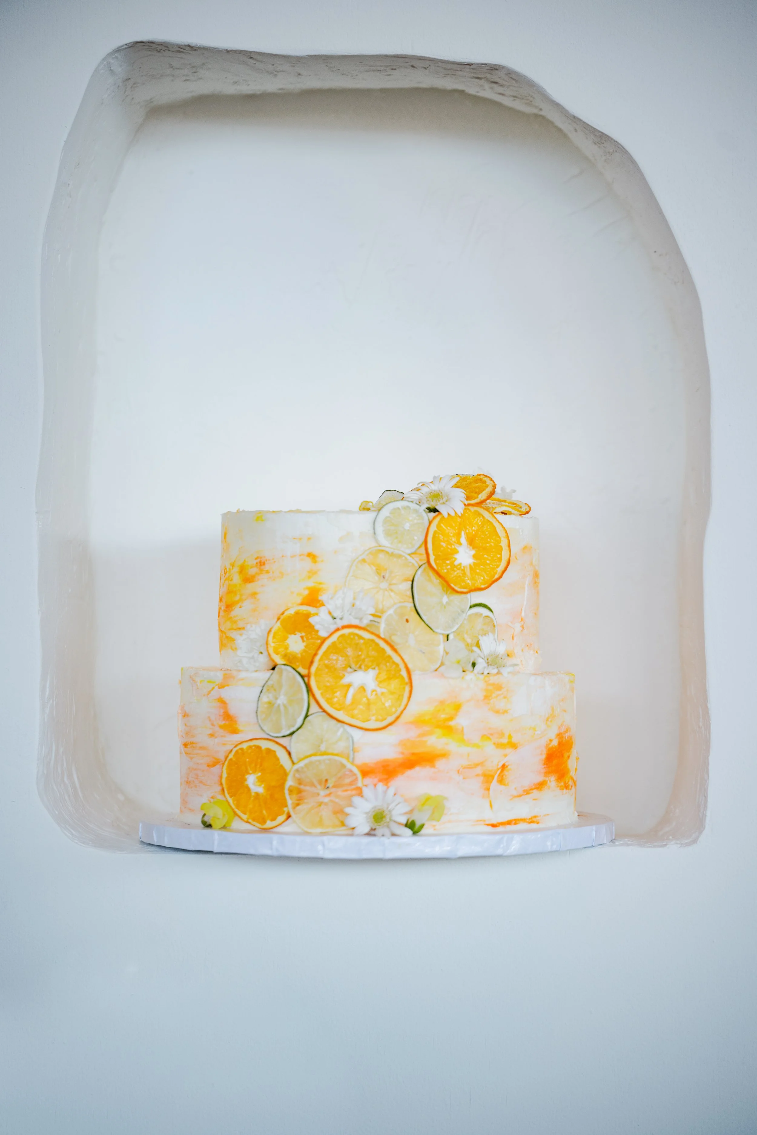 A two-tiered cake decorated with sliced oranges, lemons, cucumbers, and white flowers, displayed in a white arched niche.