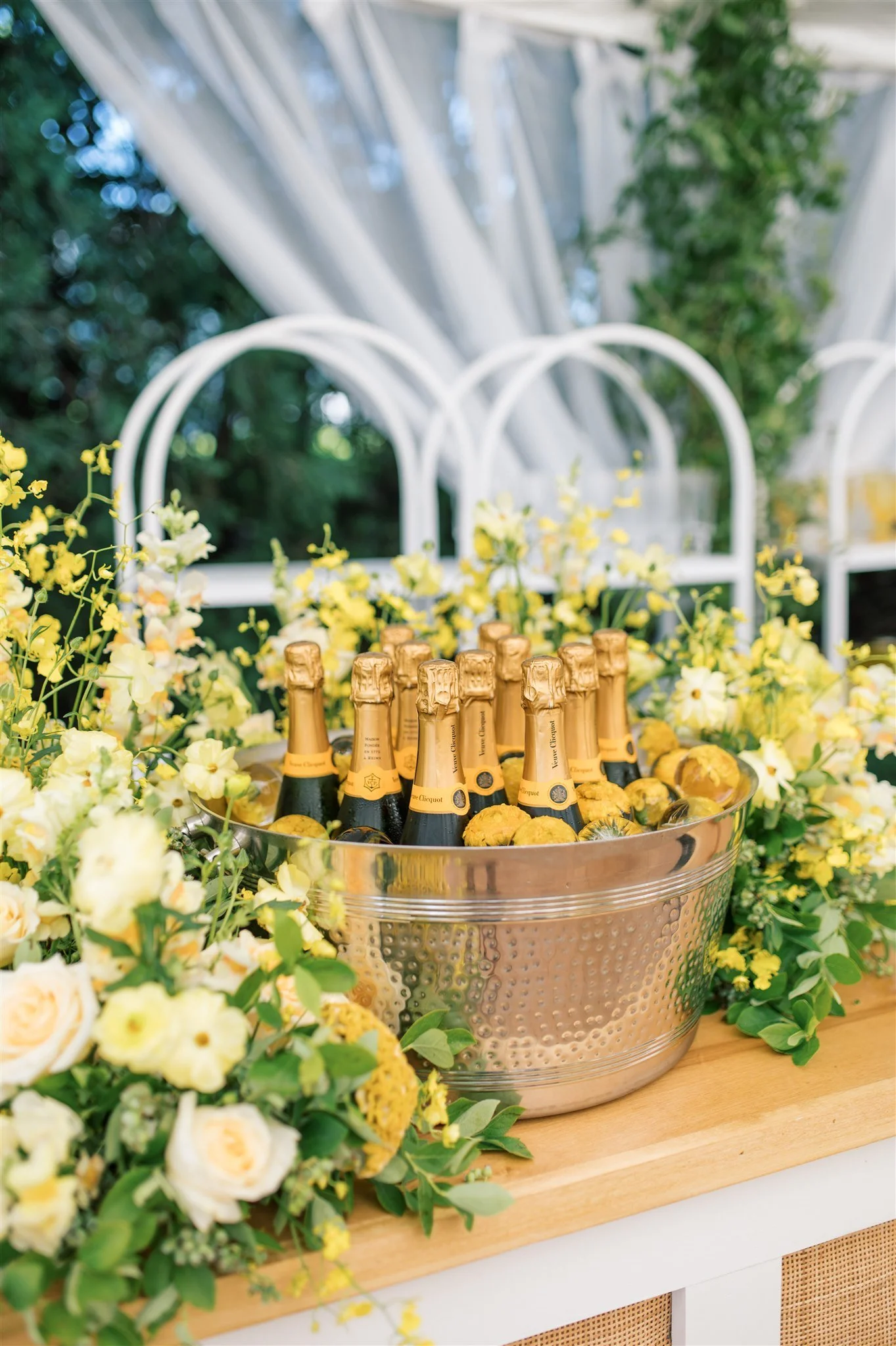 A large silver bucket filled with bottles of champagne surrounded by yellow and white flowers on a wooden table at an outdoor event.