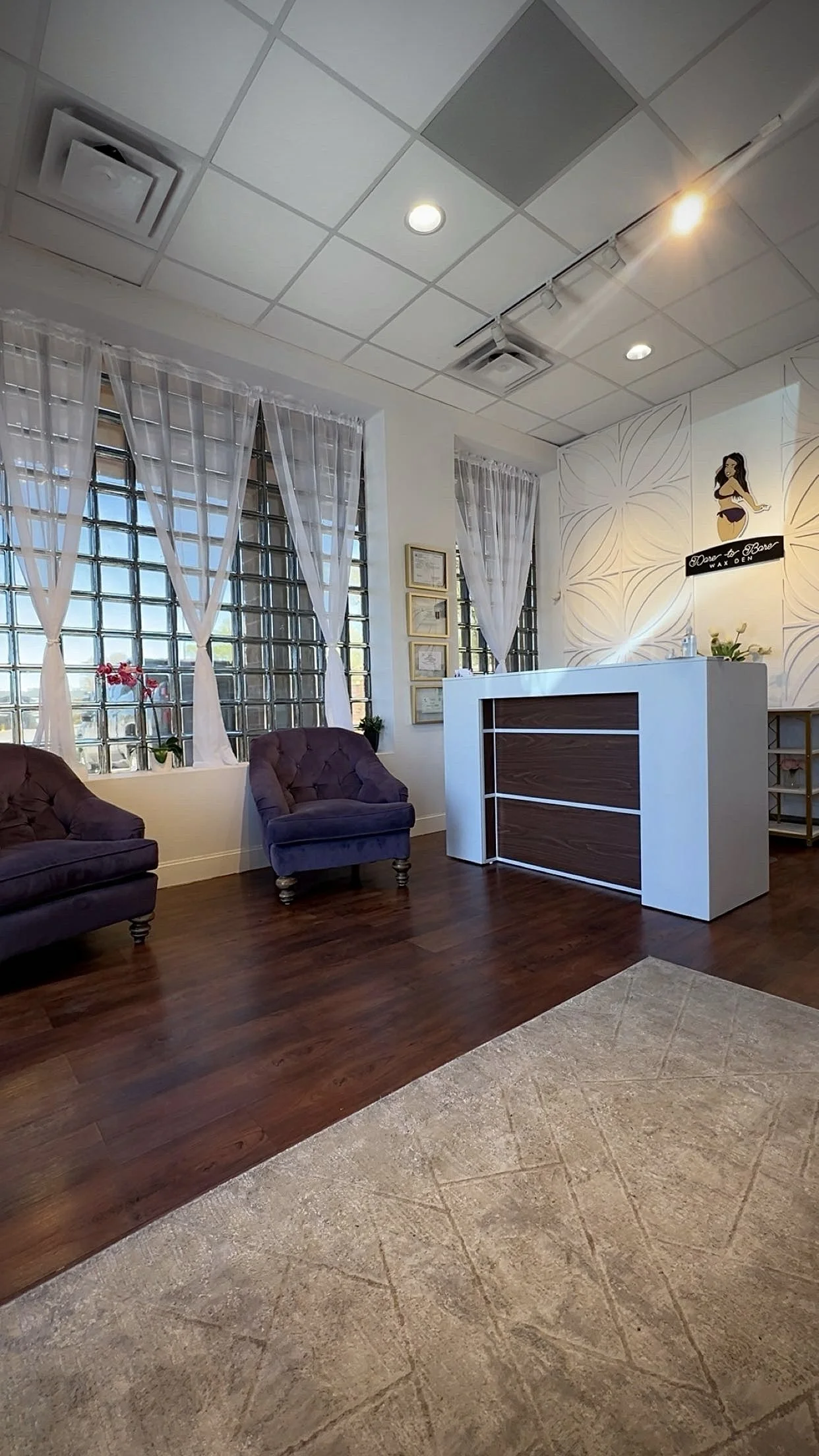Waiting room with two purple armchairs, white curtains, glass block windows, a white reception desk, framed pictures on the wall, and a sign that says "Dancer on Board" with an illustration of a woman in a dance pose.