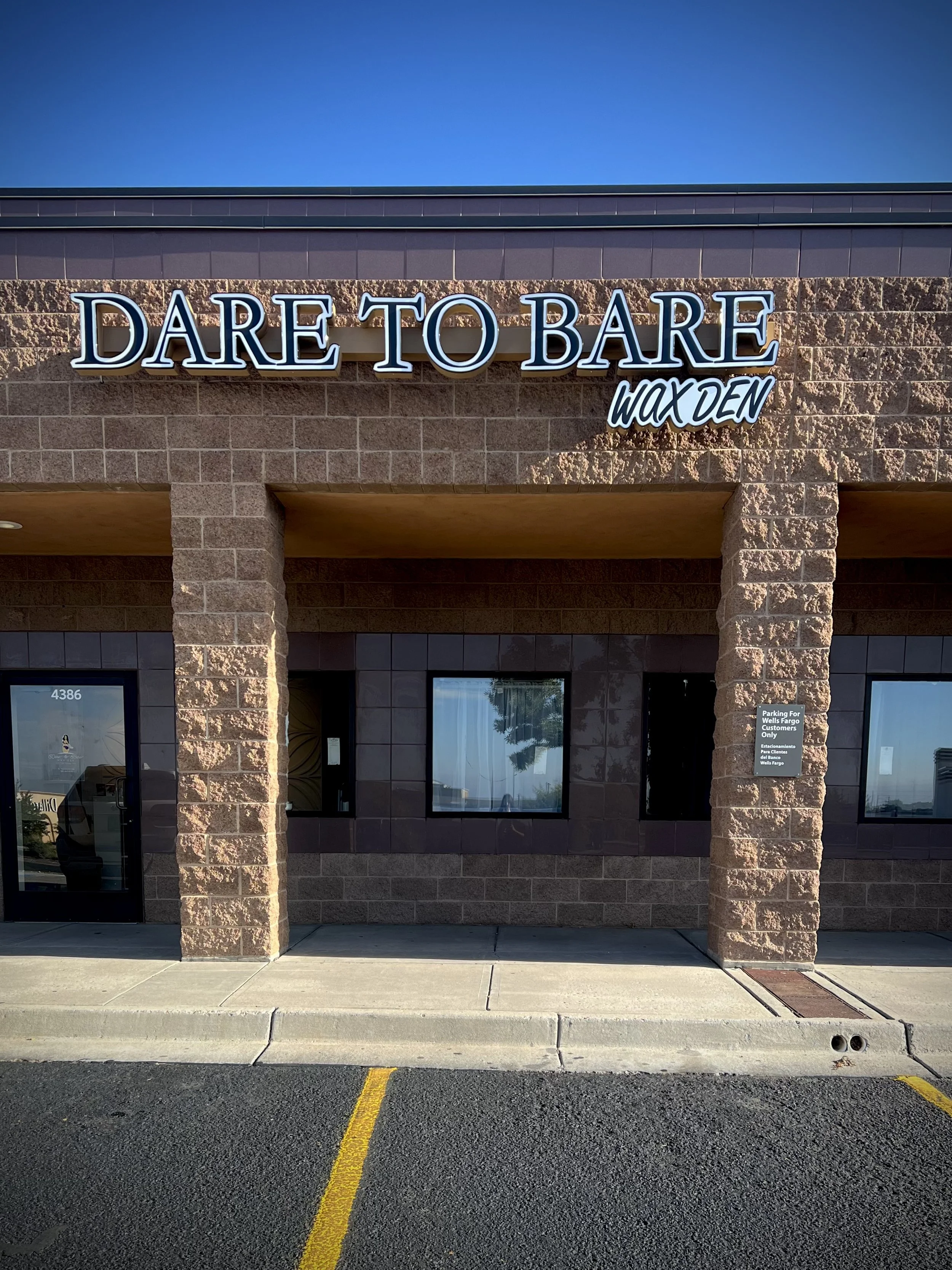 Exterior view of a storefront with a sign that reads 'DARE TO BARE WAX DEN' on a brick building, with a parking lot in front.