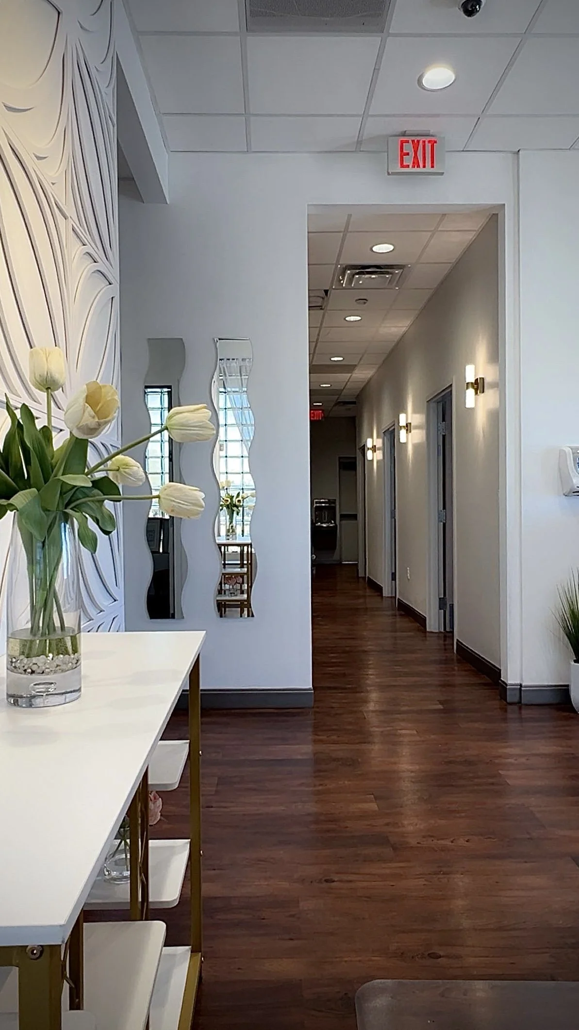 Interior view of a hallway with white walls, wood flooring, and modern lighting. There are doors along the hallway and a red exit sign above a doorway. A table with a vase of white tulips is visible on the left.