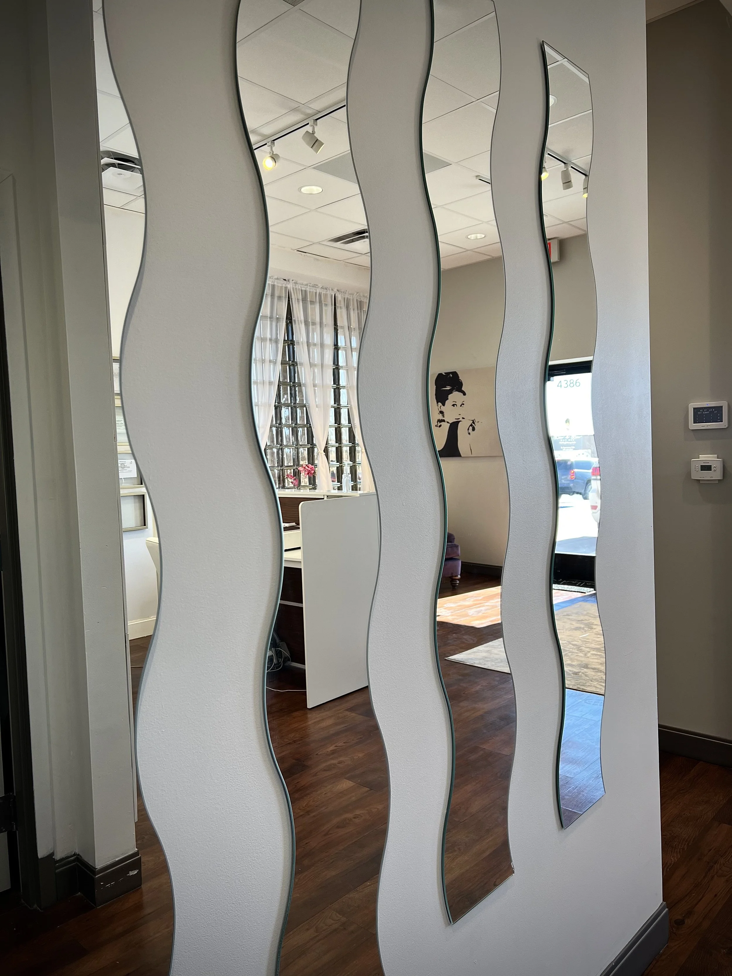 Interior view of a room with a wavy mirror wall, showing a sitting area, artwork, and large windows with curtains, wooden floors, and ceiling lights.