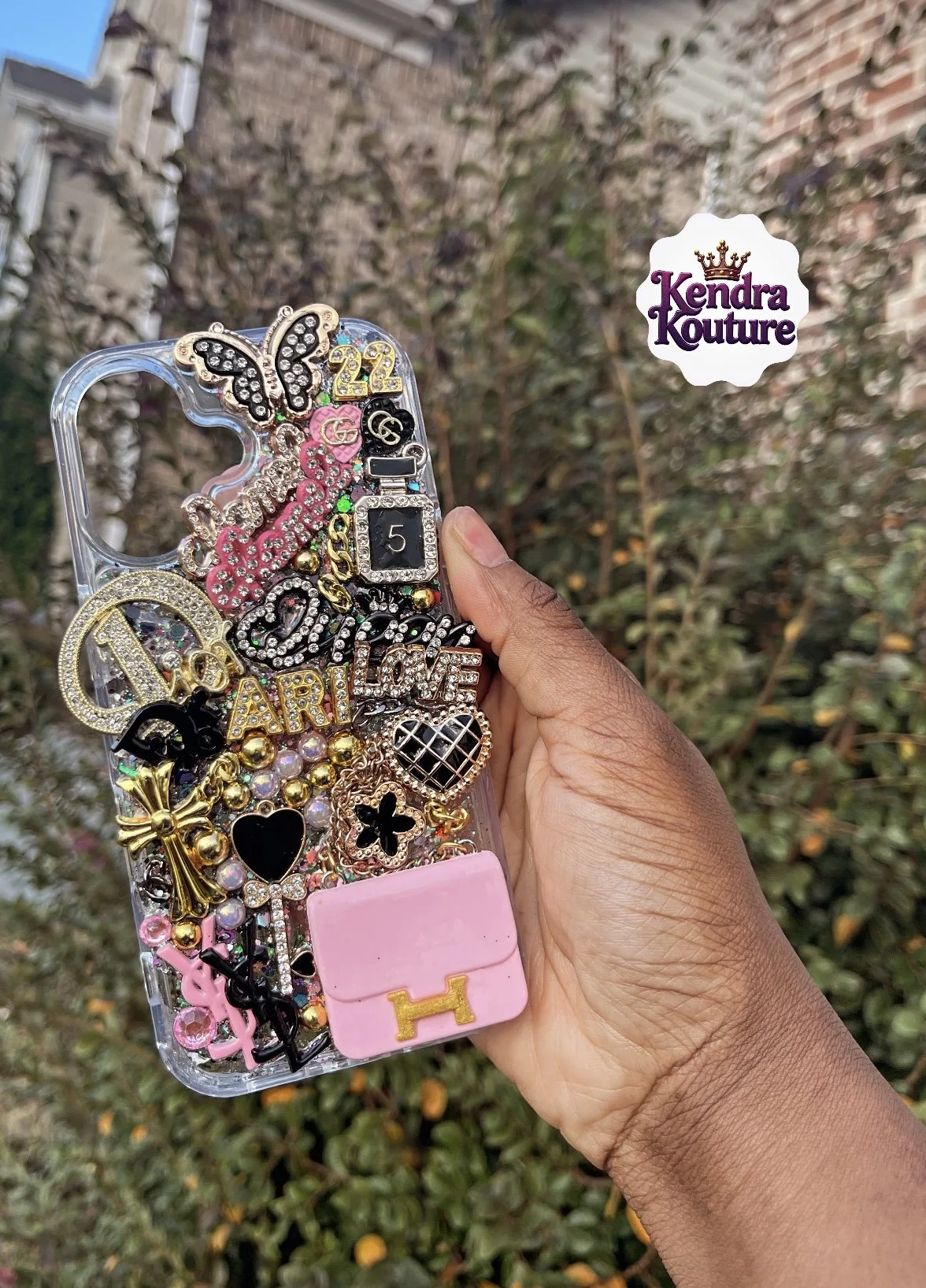 Hand holding a clear phone case decorated with various colorful and shiny jewelry and charms, including butterflies, hearts, letters, and decorative pins, against a background of trees and a brick building.