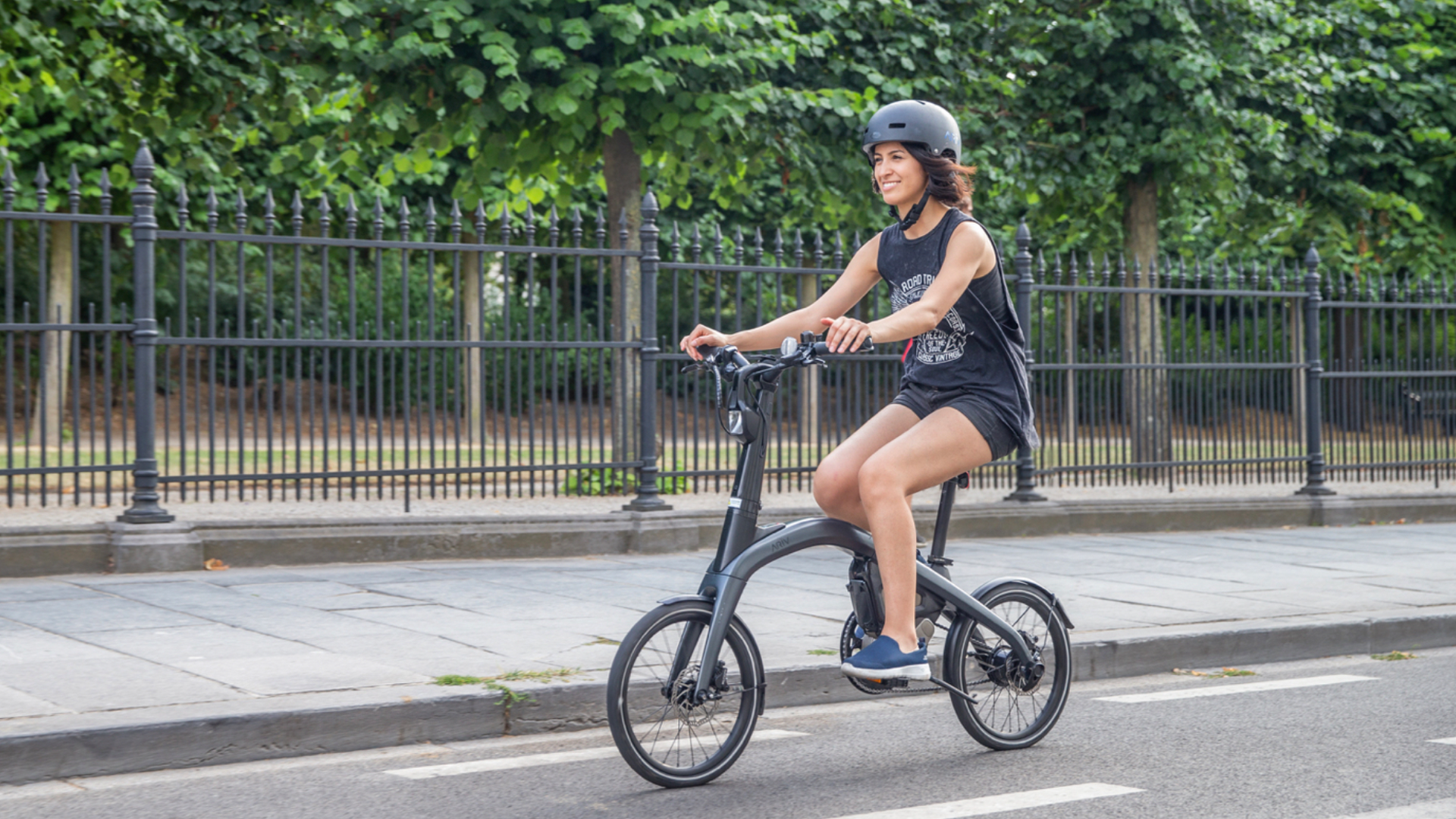 Young woman riding an electric bicycle on a city street, wearing a black helmet, sleeveless black top, shorts, and slip-on shoes, with a park and black metal fence in the background.
