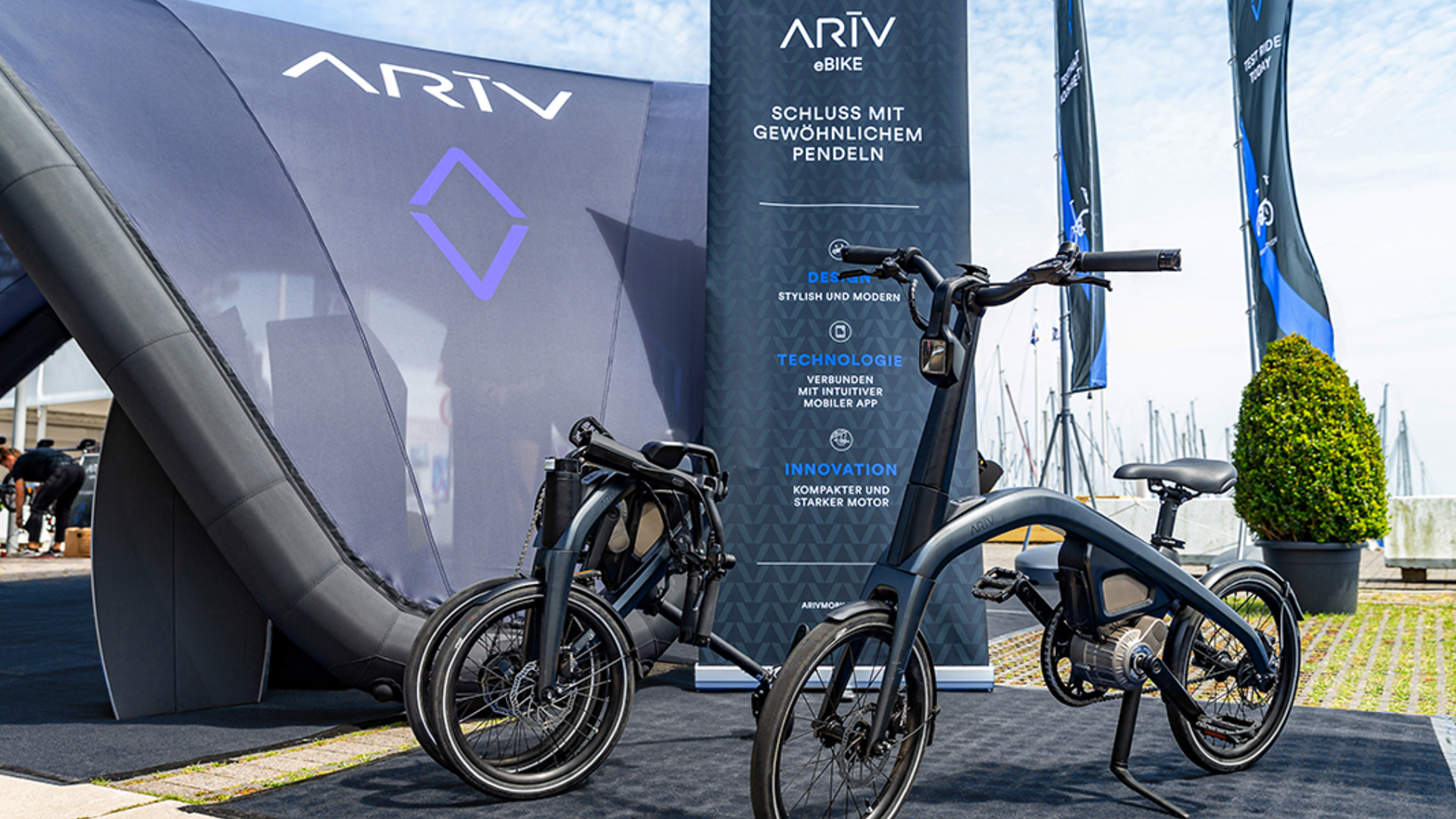 Two black electric bikes on display at an outdoor event with ARIV branding, large banners, and a marina with sailboats in the background.