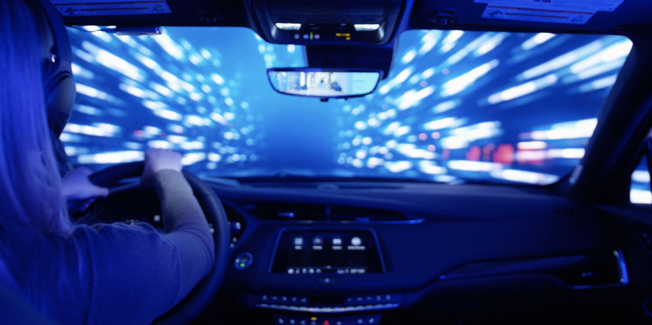 Cadillac XT4 immersive experience. Dashboard view from inside a car with blurred city lights seen through the windshield, person with light-colored hair driving.