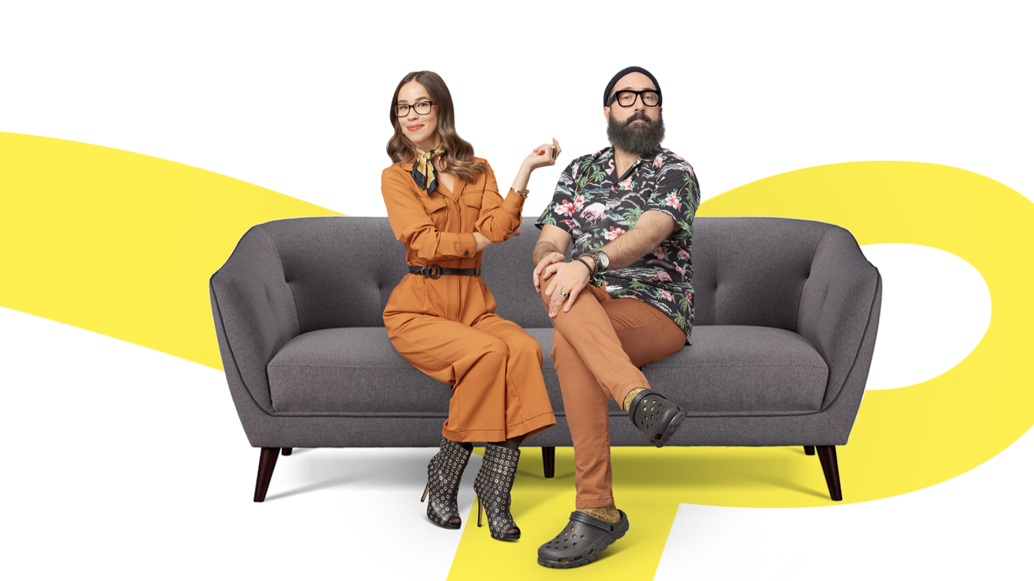 A woman and a man sitting on a gray couch against a background with yellow and white abstract shapes. The woman wears an orange jumpsuit, glasses, and patterned high heels. The man wears glasses, a floral shirt, brown pants, and black Crocs.