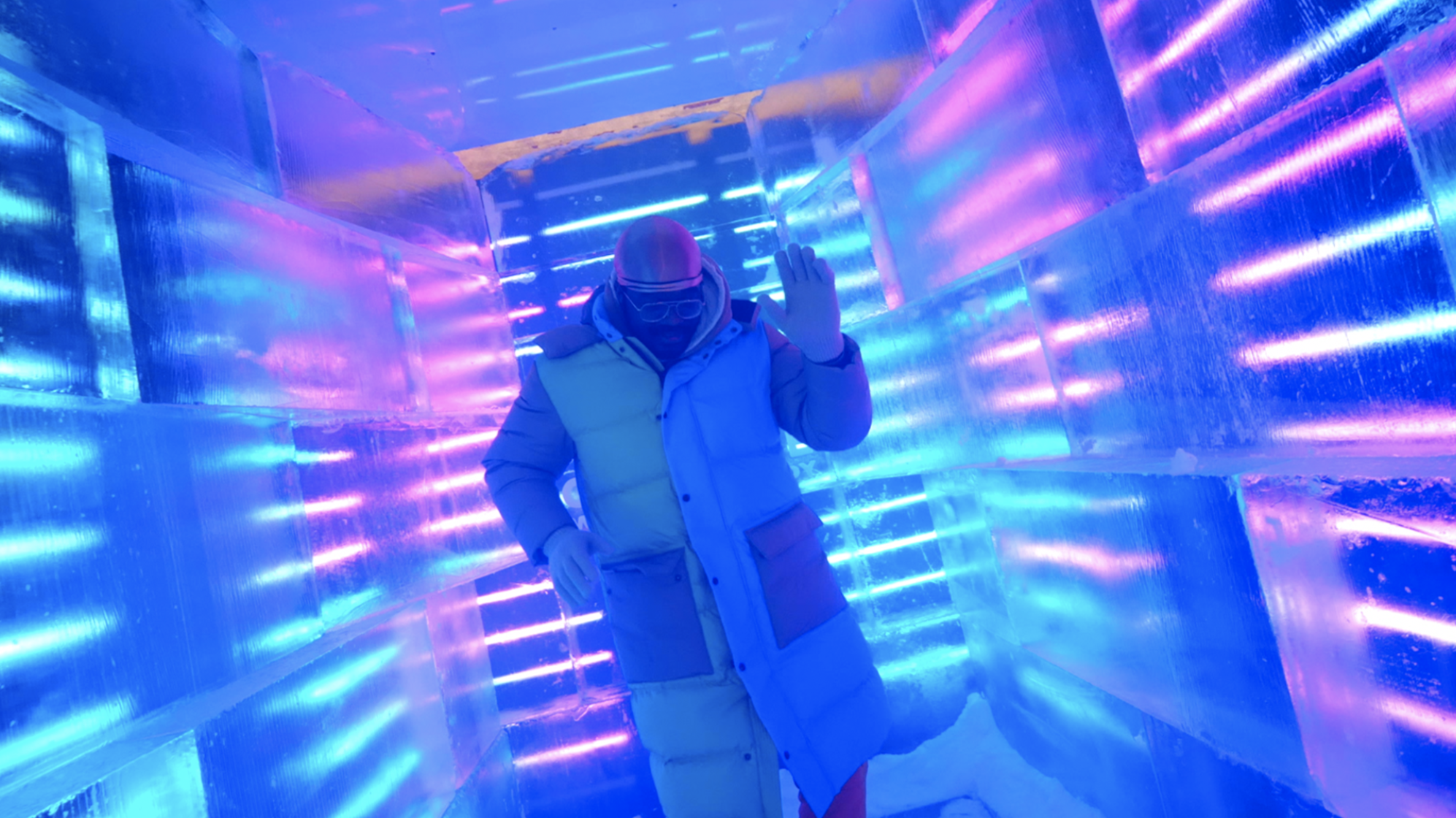 Person in winter clothing, wearing a mask and glasses, standing in an ice tunnel illuminated with colorful blue, purple, and pink neon lights, waving with one hand.