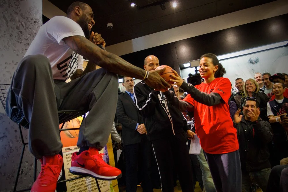 Lebron James is passing a basketball to a smiling young girl in a red shirt during an event, with a crowd of spectators in the background.