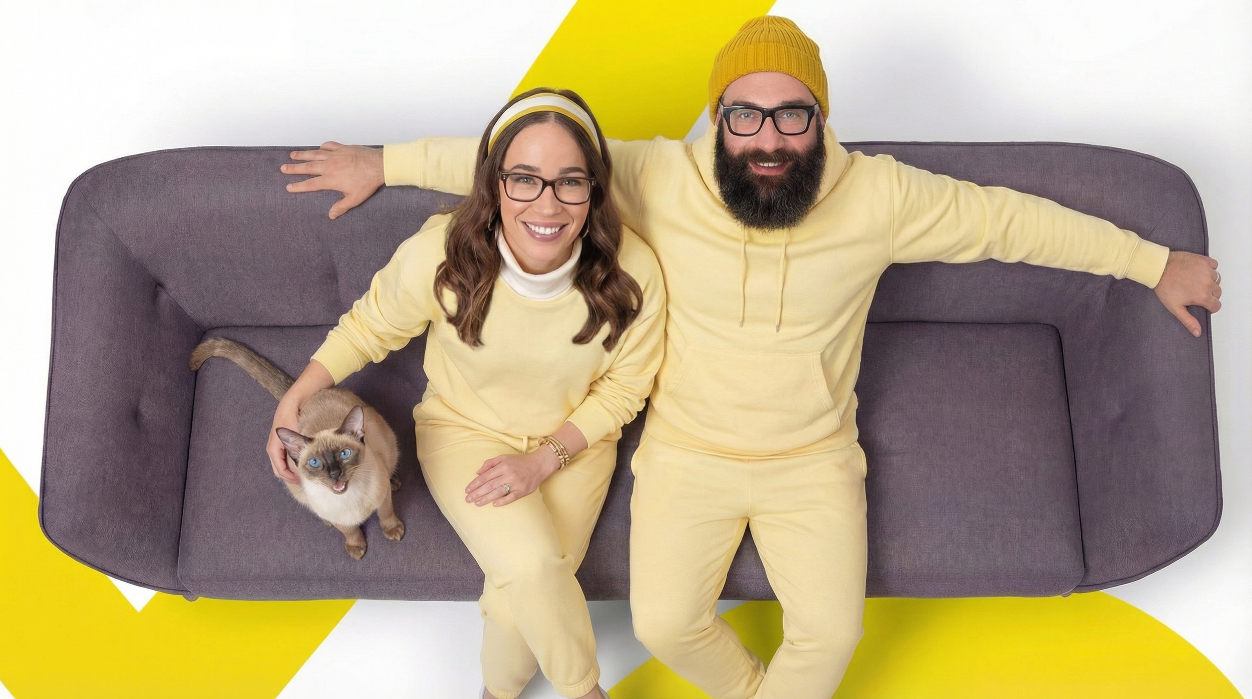 A happy couple and a cat sitting on a gray couch, all wearing yellow clothes. The woman has glasses and a headband, the man has glasses and a yellow beanie, and the woman is petting a Siamese cat with blue eyes.