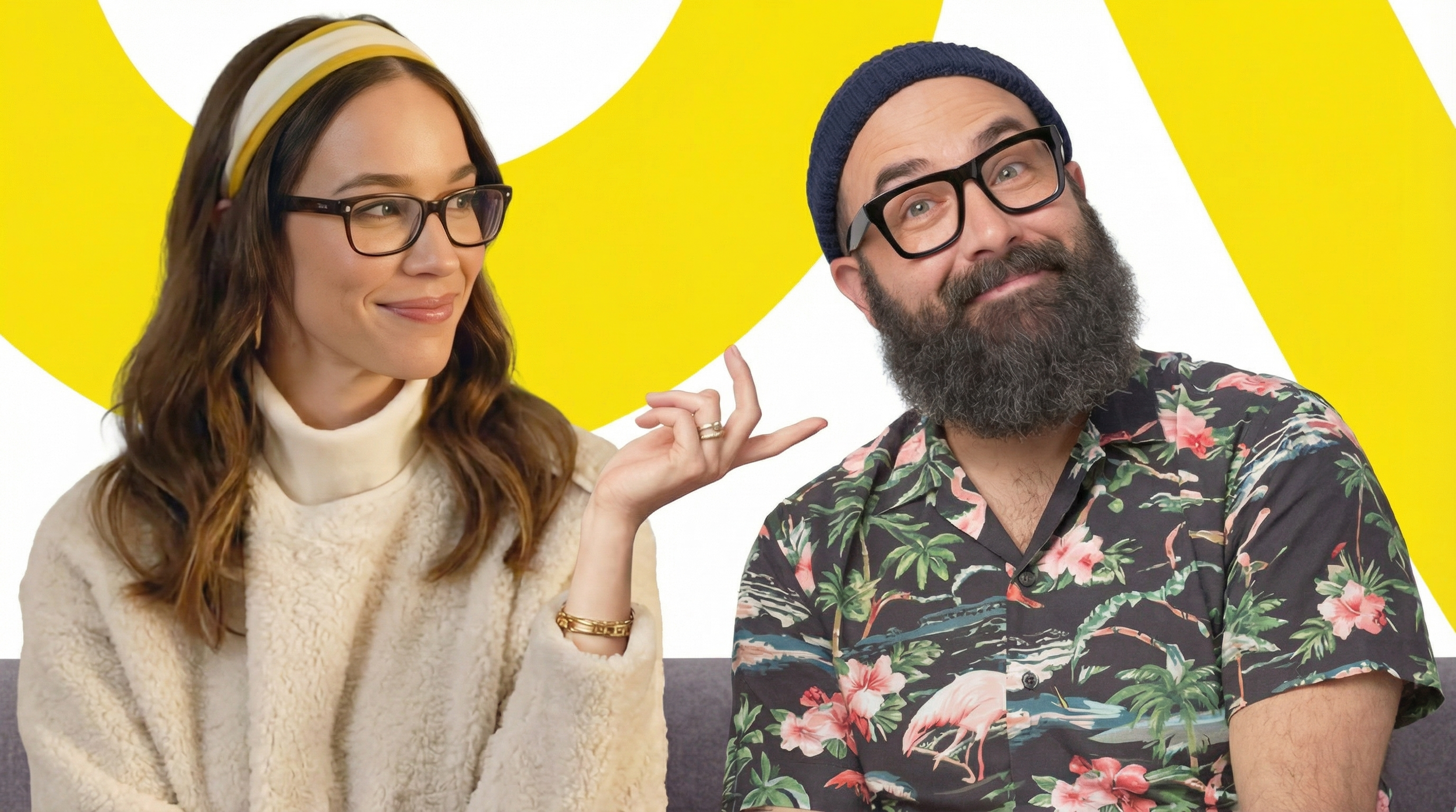 Woman with glasses and brown hair wearing a cream turtleneck and headband, smiling and pointing at man with glasses and a beard, wearing a Hawaiian shirt and navy beanie, sitting on a couch with a yellow and white abstract background.