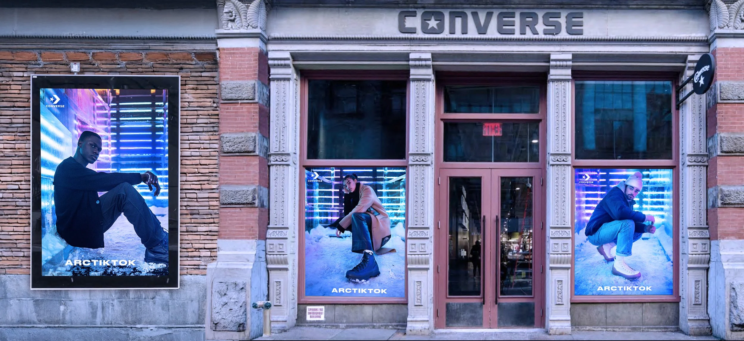 Exterior of a Converse store with large digital posters of models in winter clothing. The posters feature models in snowy environments with neon lighting, and the store has a brick facade with decorative stone accents.