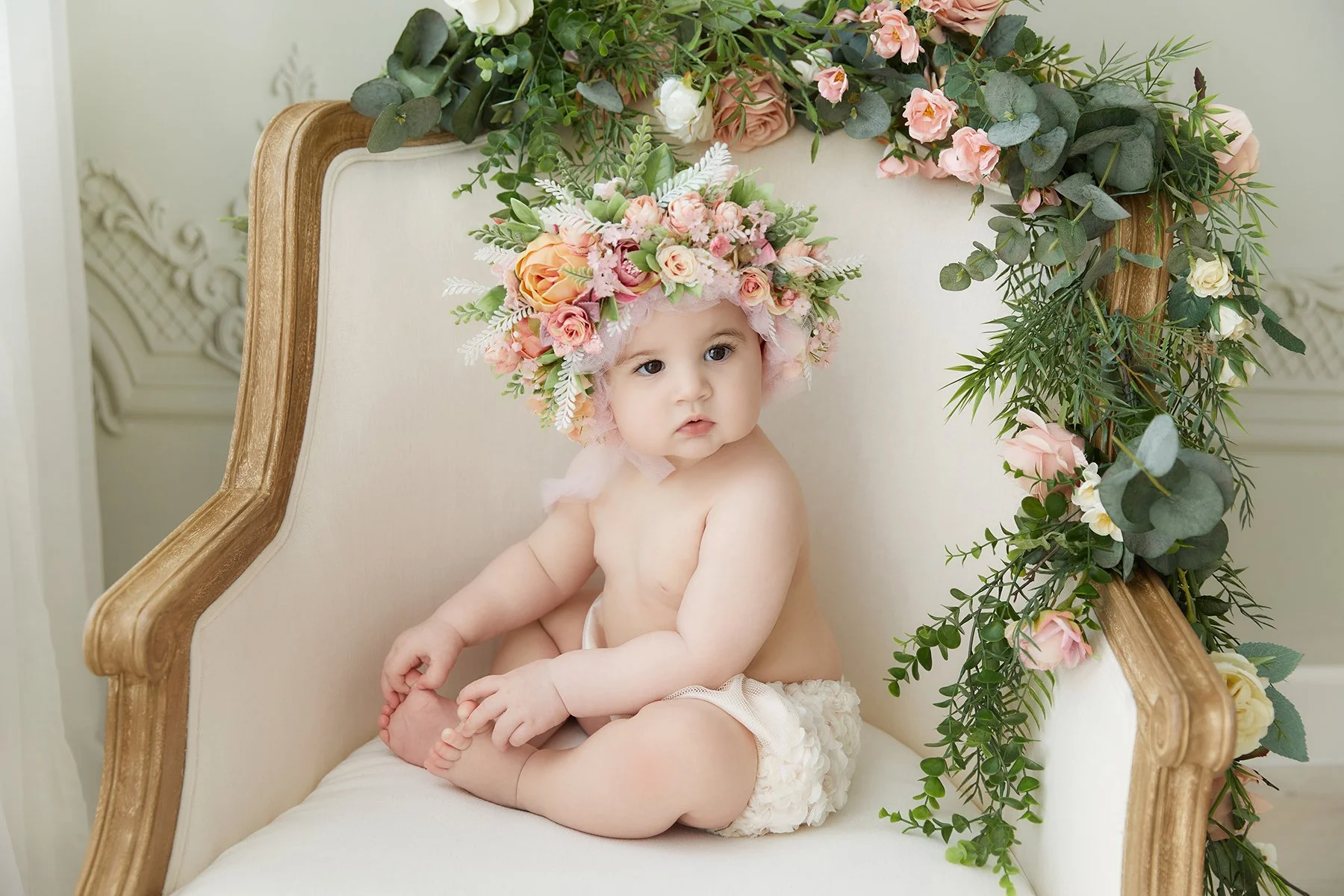 A baby sitting on an elegant cream-colored chair decorated with pink, peach, and white flowers and green foliage, wearing a floral crown.