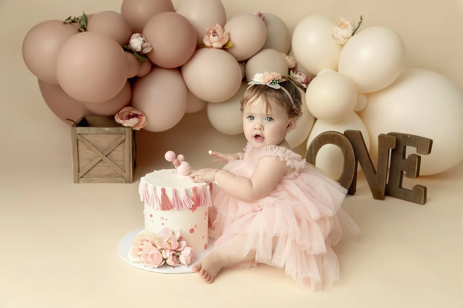 A young girl in a pink tutu dress sits next to a white birthday cake decorated with pink tassels and flowers, with a backdrop of pink and cream balloons and a wooden 