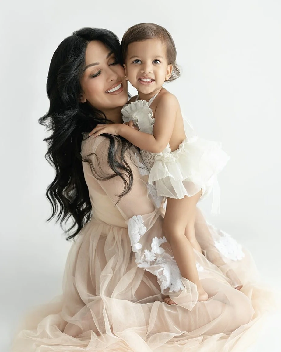 A woman with long dark hair holding a smiling young girl, both wearing white dresses, in front of a plain white background.
