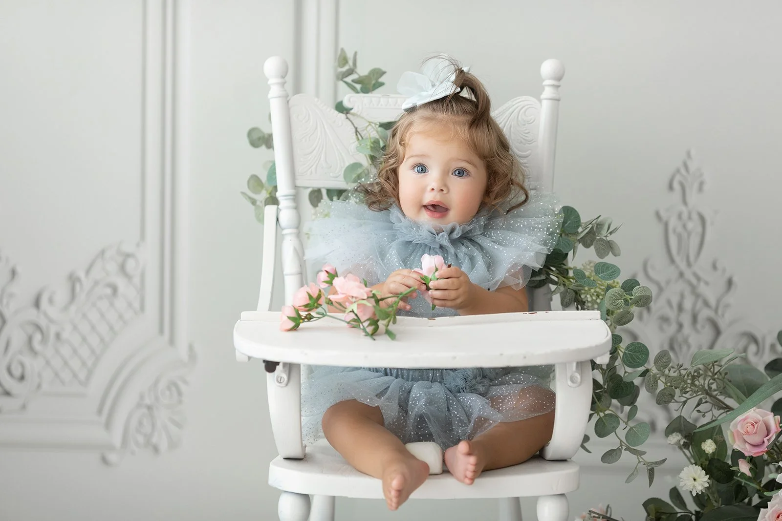 A young girl with curly hair, blue eyes, and wearing a sparkly blue tulle dress with a large white bow in her hair, sitting in a white high chair surrounded by pink and white flowers and greenery, in a bright room with white walls and decorative mold