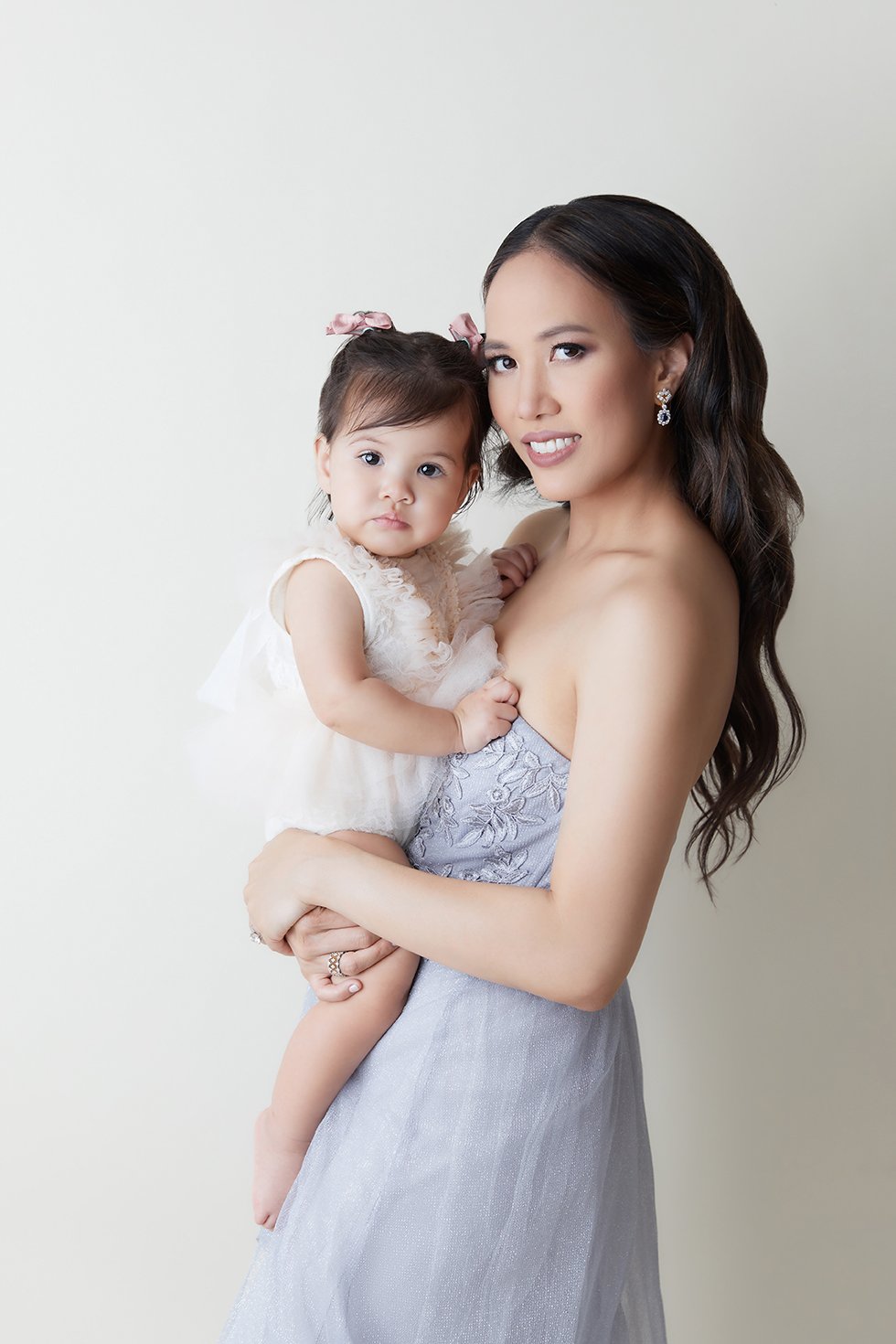 A woman in a strapless dress holding a young girl in a white dress against a plain background.