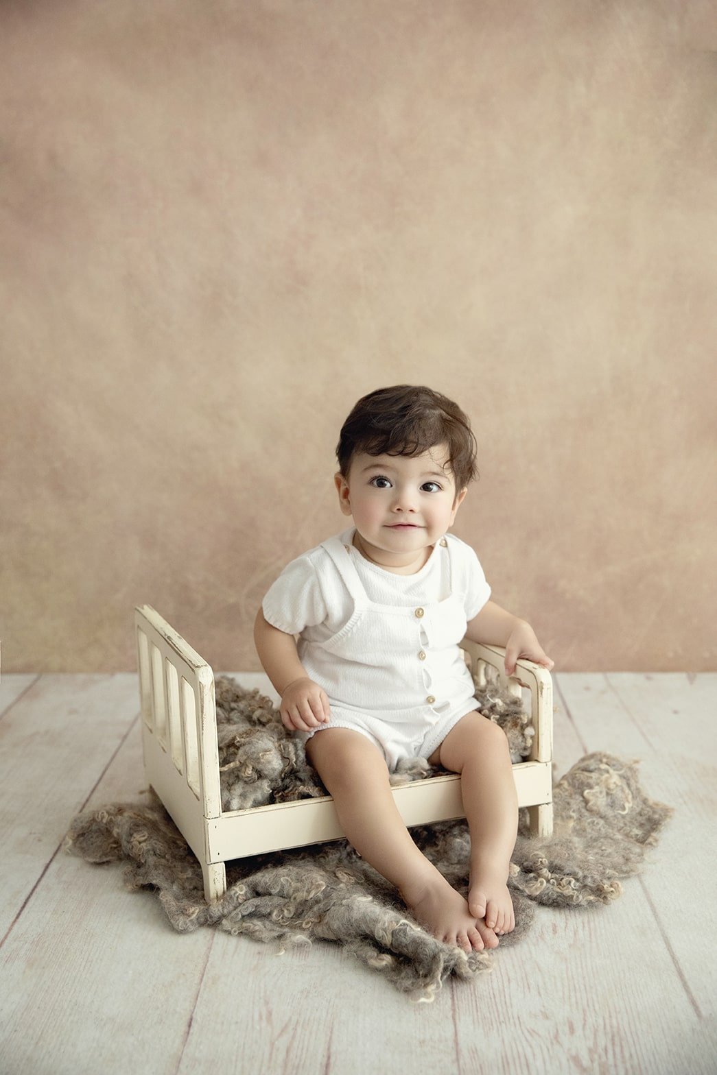 A young child sitting on a small bed with a fluffy blanket, dressed in a white romper, against a plain beige background.