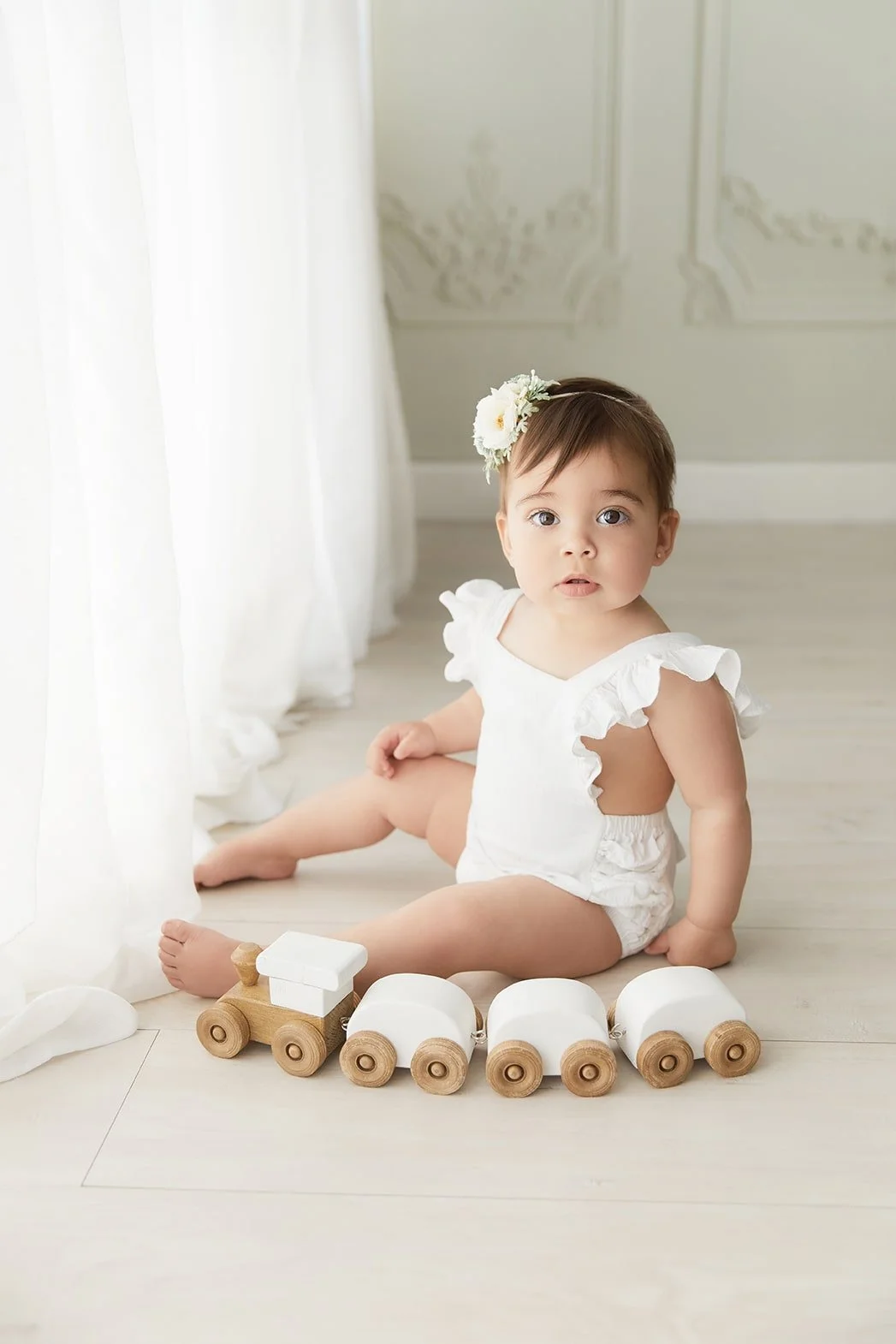A young girl sitting on a light wooden floor near a white curtain, dressed in a white outfit with ruffled sleeves, with a white flower headband, and playing with a wooden and white toy train.