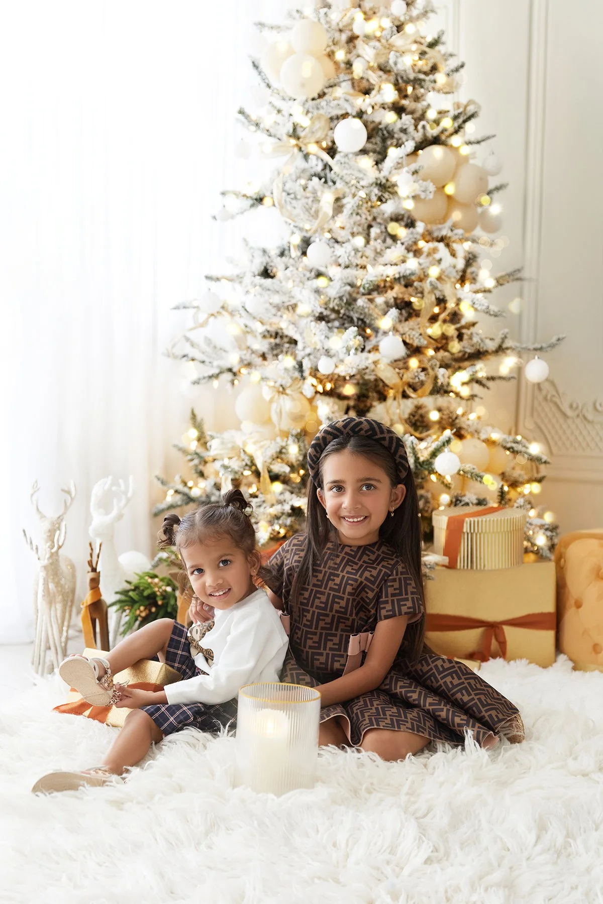 Two young girls sitting on a white fluffy rug in front of a decorated Christmas tree with white ornaments and lights, surrounded by wrapped presents and holiday decorations.