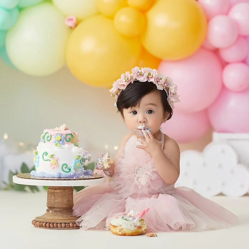 A young girl in a pink dress and floral crown sitting on the floor with a cake and balloon backdrop at a birthday celebration.