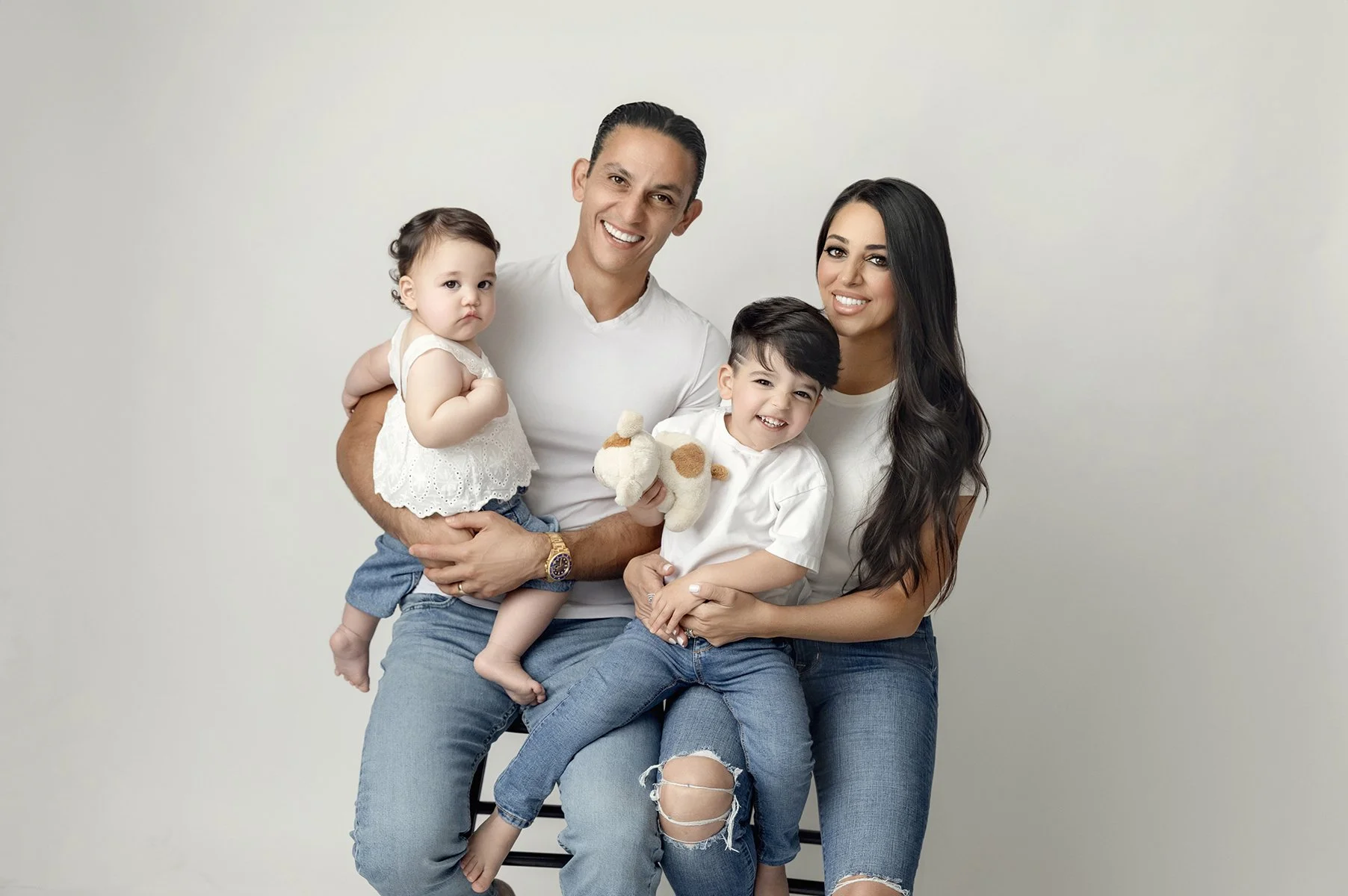 A happy family of four posing together against a plain background. The father, mother, and their two young children, a boy and a girl. The father is holding the girl on his lap, the mother has her arm around the boy, and they all are smiling.