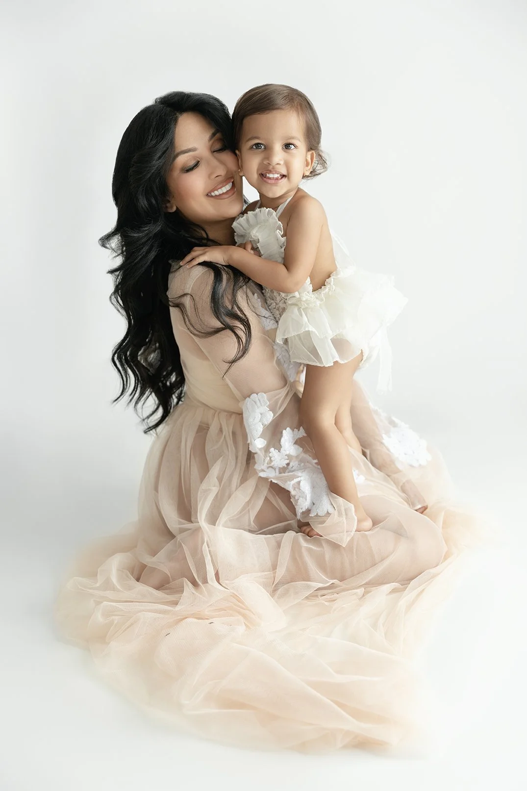 A woman with long, dark hair and a young girl with brown hair smiling and hugging each other. The woman is kneeling on a white surface, wearing a flowing, peach-colored dress. The girl is wearing a white dress with ruffles.
