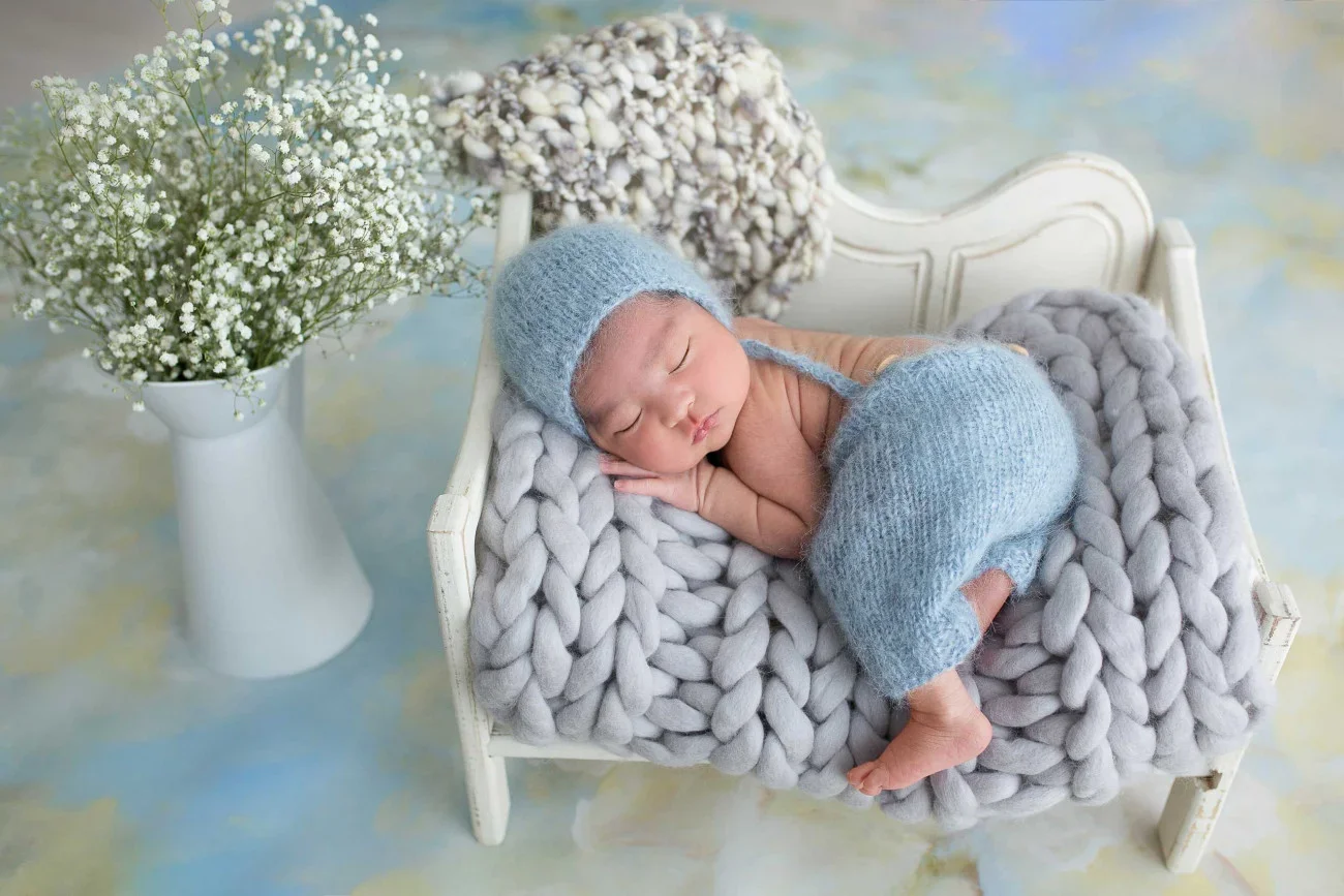 A sleeping newborn baby lying on a small white bed with a chunky gray knit blanket, wearing a matching blue knit hat and diaper cover, next to a white vase filled with small white flowers.