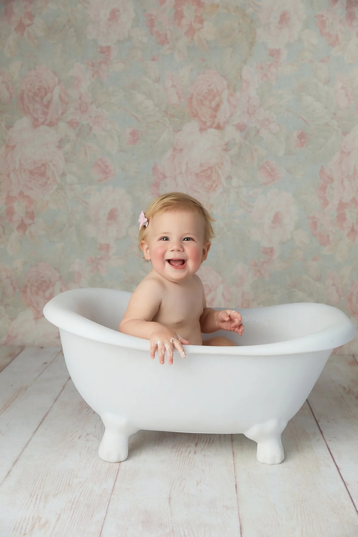 A happy baby sitting in a small white bathtub with a floral background.