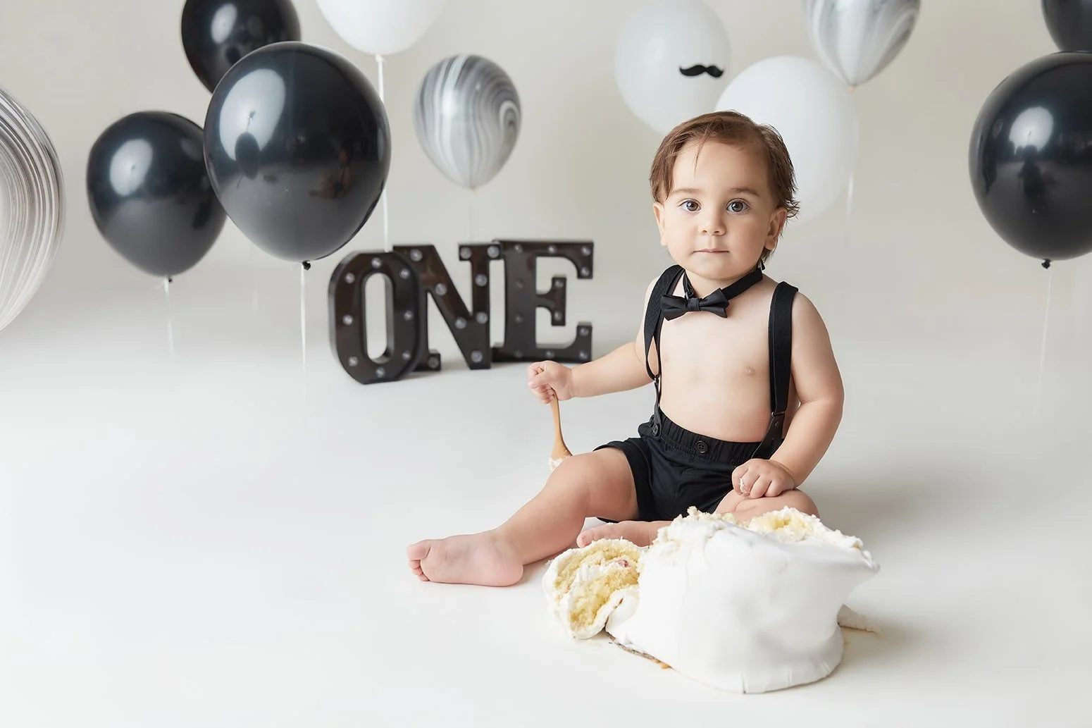 Baby boy sitting on the floor with birthday cake in front, surrounded by black, white, and marbled balloons, with a black "ONE" sign in the background.